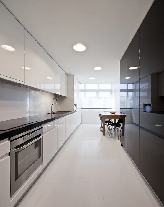 A minimalist galley kitchen with high-gloss white and black cabinets, reflective flooring, and a small dining table by a bright window.