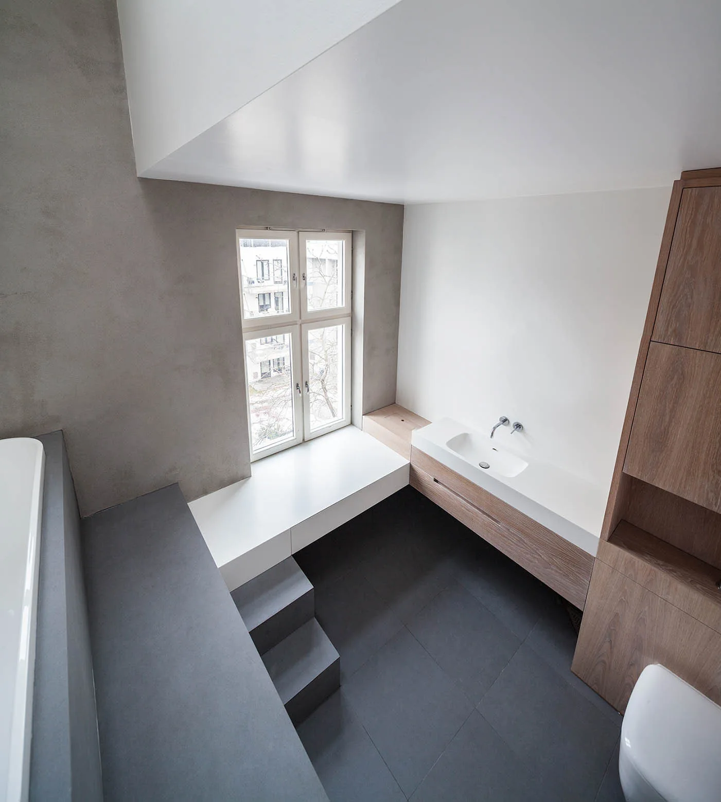 Minimalist bathroom with a sunken dark grey tiled floor, small steps, a floating wood vanity, and a white built-in window bench.