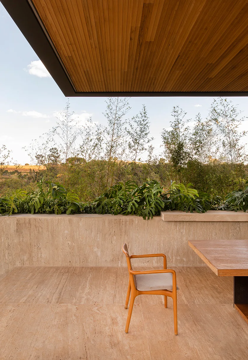 An outdoor patio with travertine stone flooring and walls, a slatted wood ceiling, a wooden dining chair, and leafy green plants in the background.