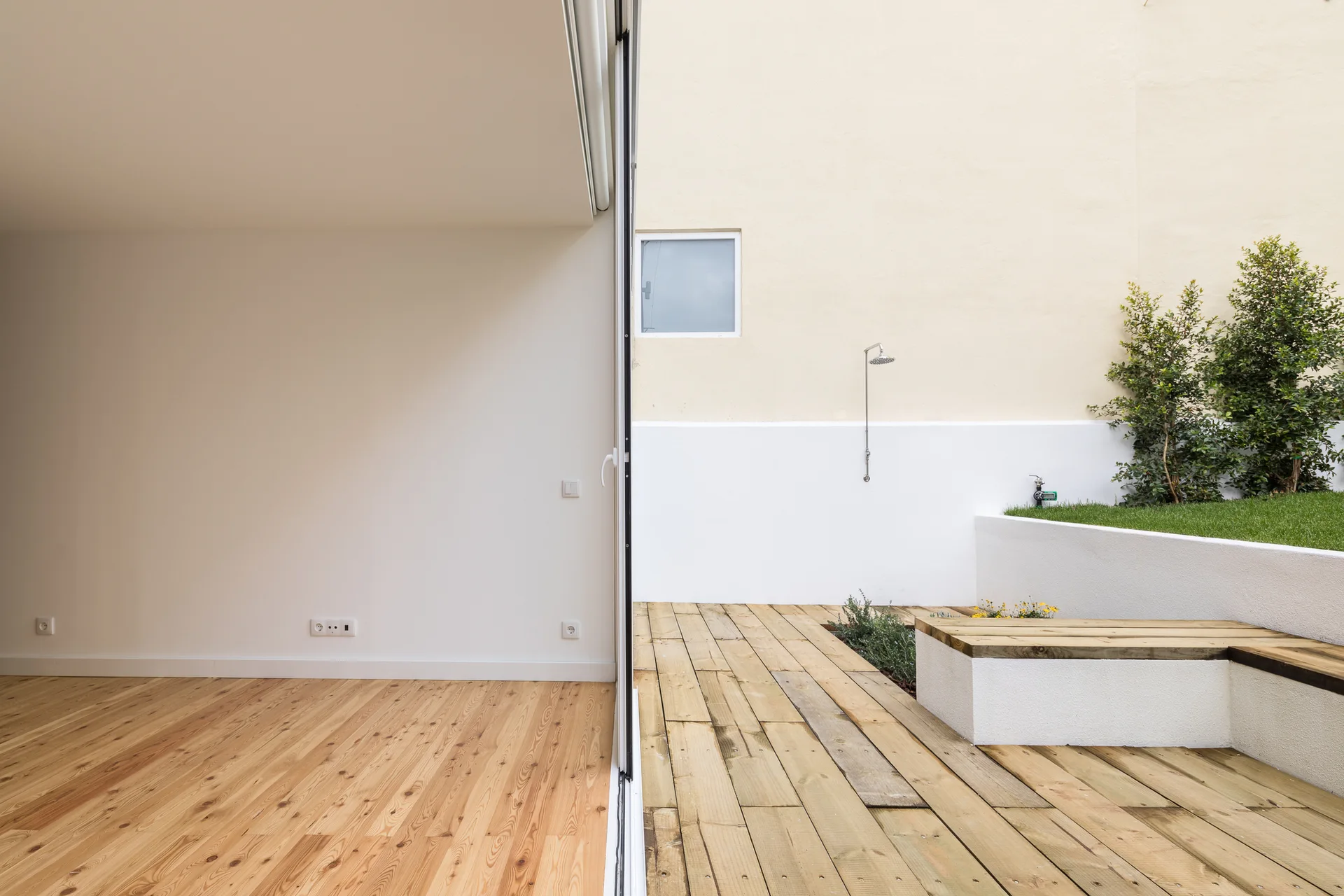 A minimalist indoor room with wood floors opens onto a wooden patio deck featuring a built-in bench and wall-mounted outdoor shower.