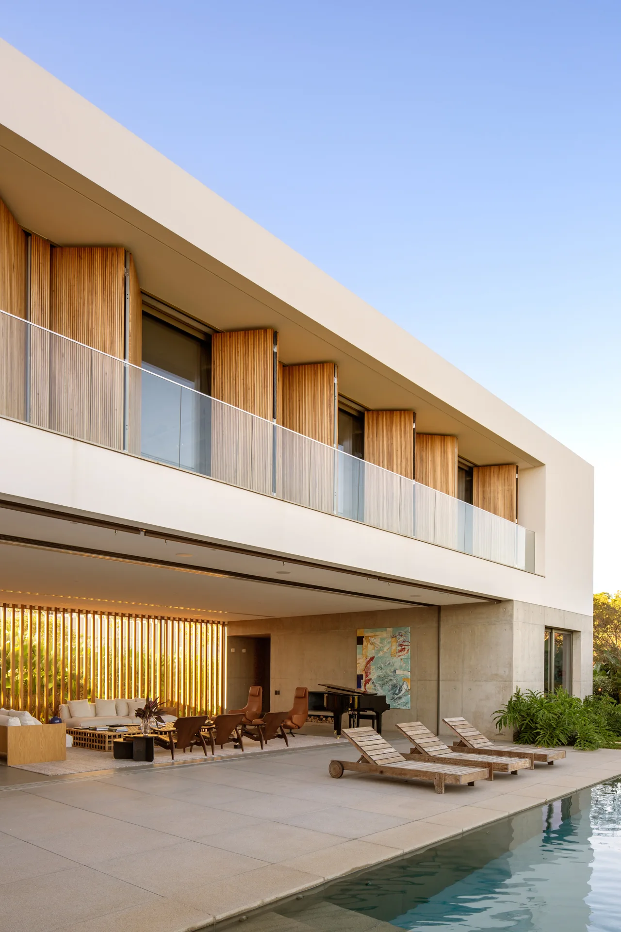 Three wooden lounge chairs sit beside a swimming pool next to an open-plan living area with concrete walls and a vertical wood slat screen.