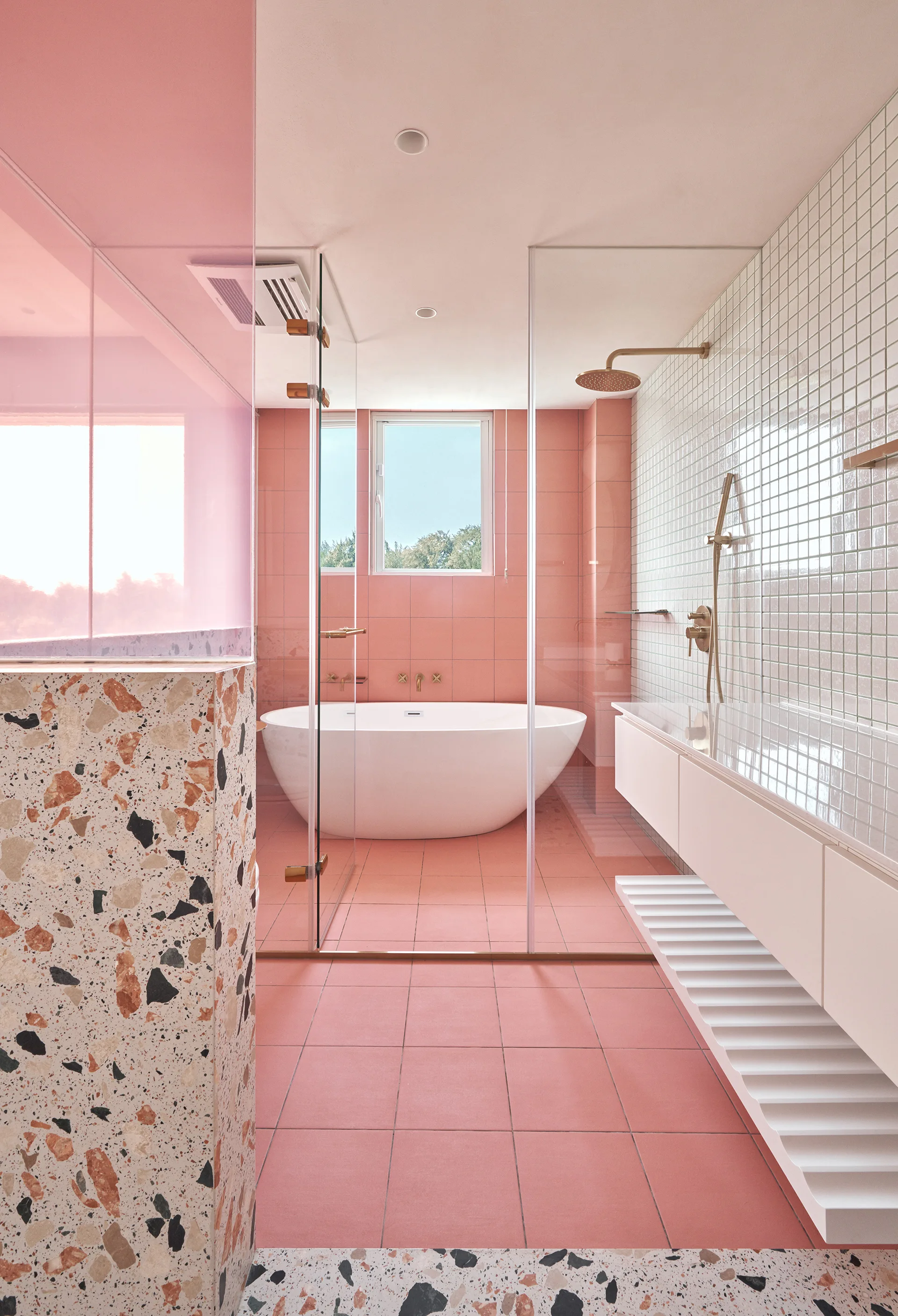 A modern pink-tiled bathroom features a freestanding white bathtub, a glass-enclosed shower, brass fixtures, and a prominent speckled terrazzo partition.