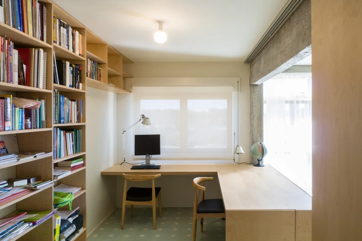 A minimalist home office featuring an L-shaped light wood desk, built-in bookshelves, and two wooden chairs beneath an exposed concrete beam.