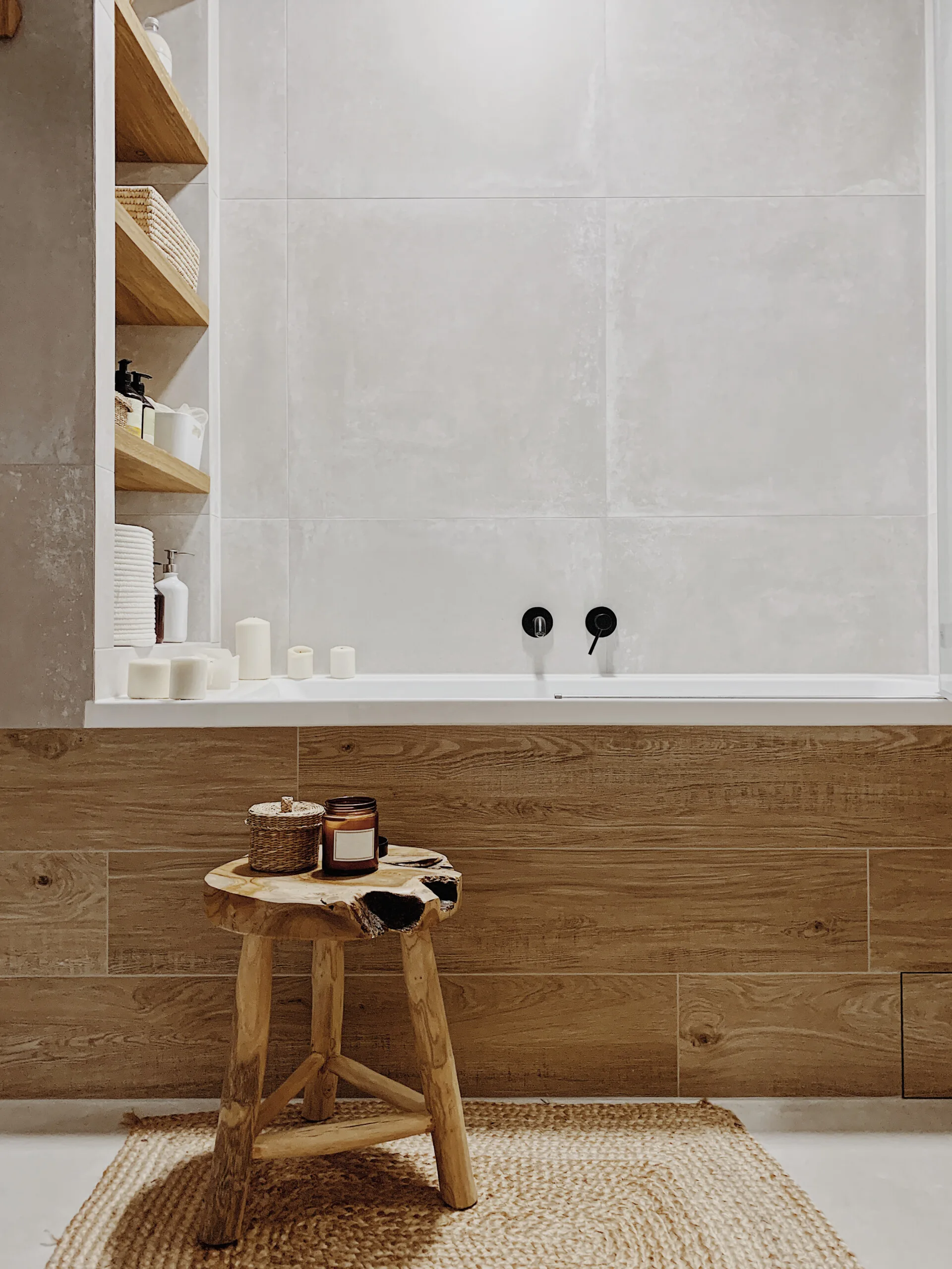 A modern bathroom features a white bathtub clad in wood-grain tile, a natural wood stool on a woven rug, and recessed wall shelves.