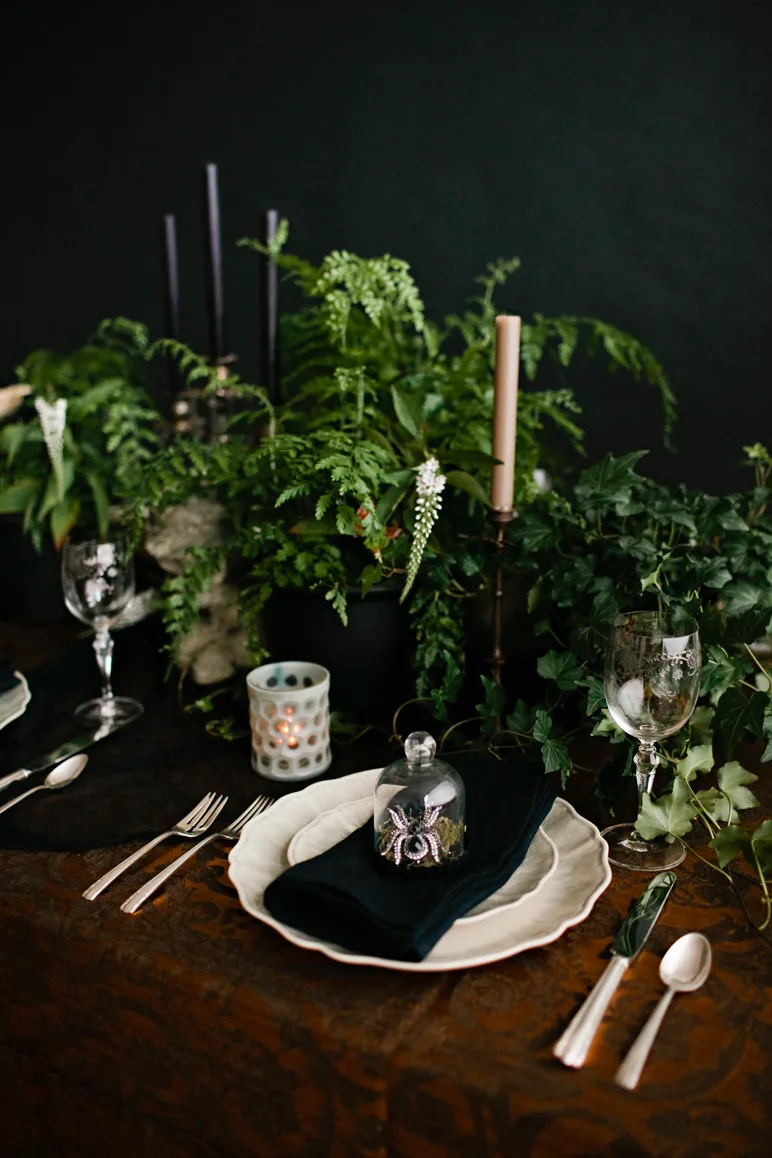A moody dining table setting features a scalloped white plate, a black napkin under a small glass cloche with a decorative spider, and lush green ferns.