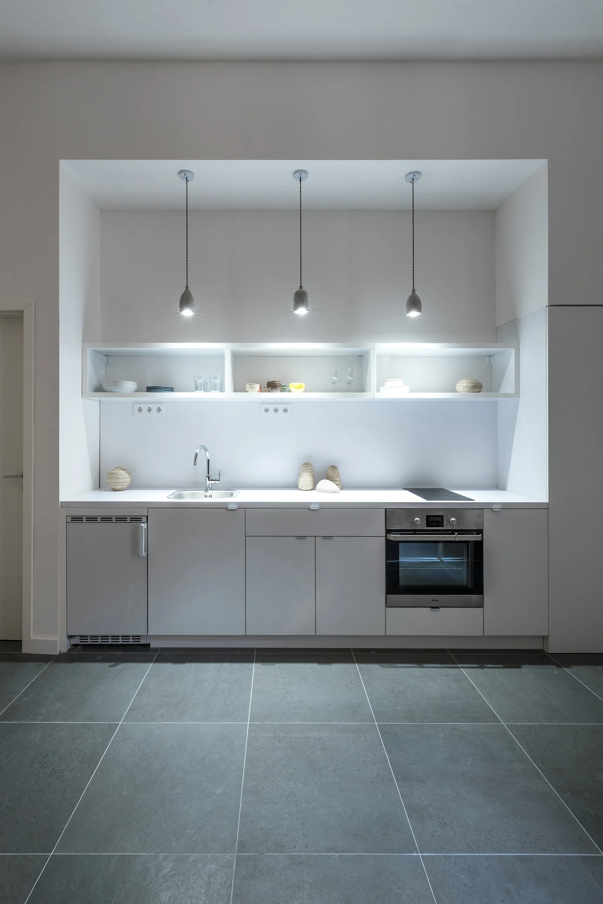A minimalist grey kitchen built into a recessed white alcove features open shelving, three pendant lights, and a dark grey tiled floor.