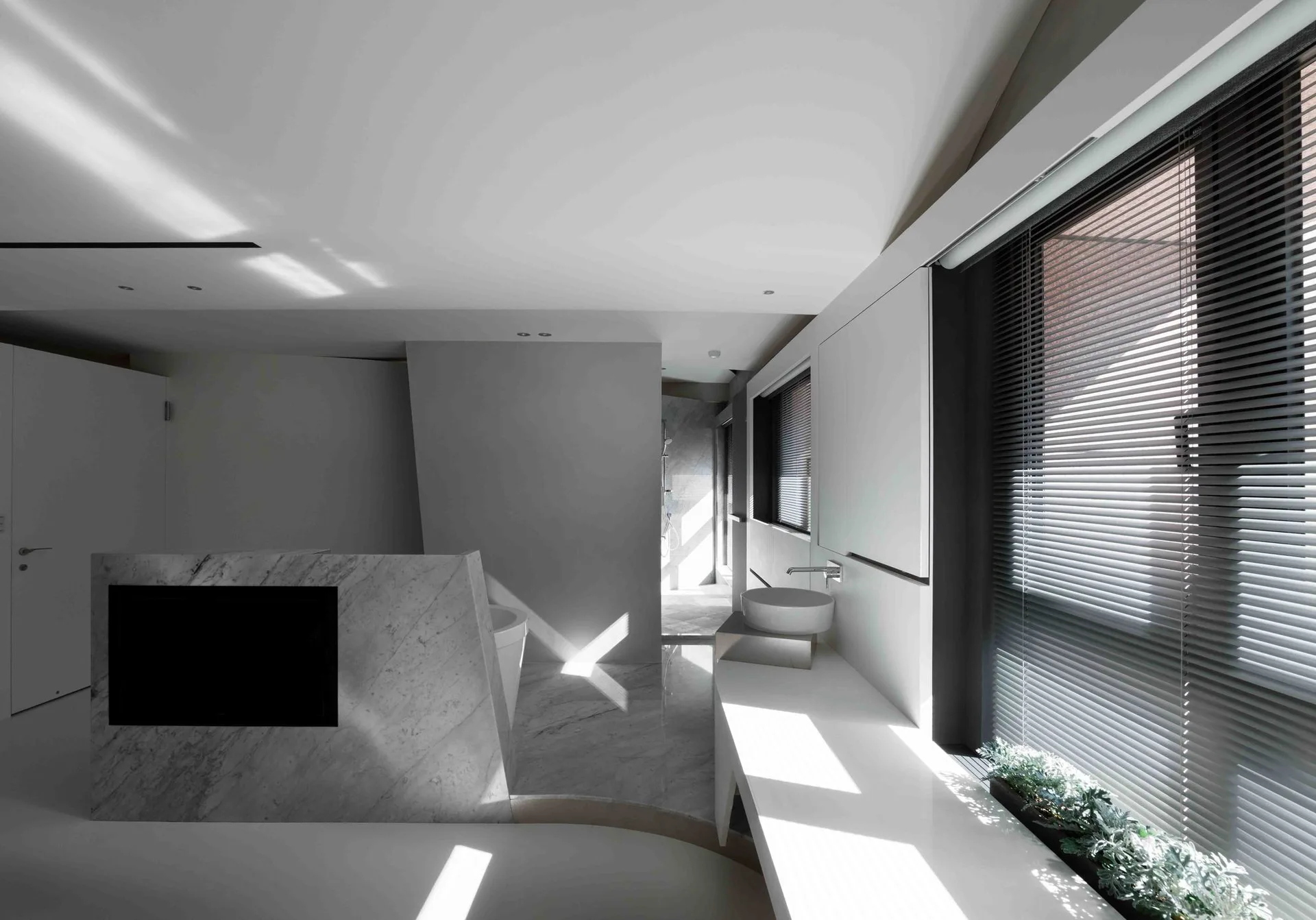A modern minimalist bathroom features a white built-in vanity with a circular vessel sink, a marble-clad partition wall, and horizontal window blinds.