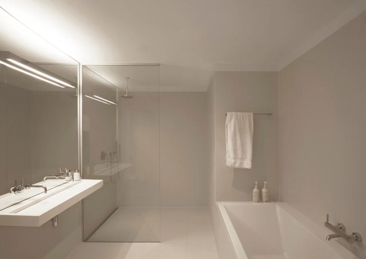 A modern minimalist bathroom with light grey tiled walls, a white floating trough vanity, a glass shower partition, and a built-in bathtub.