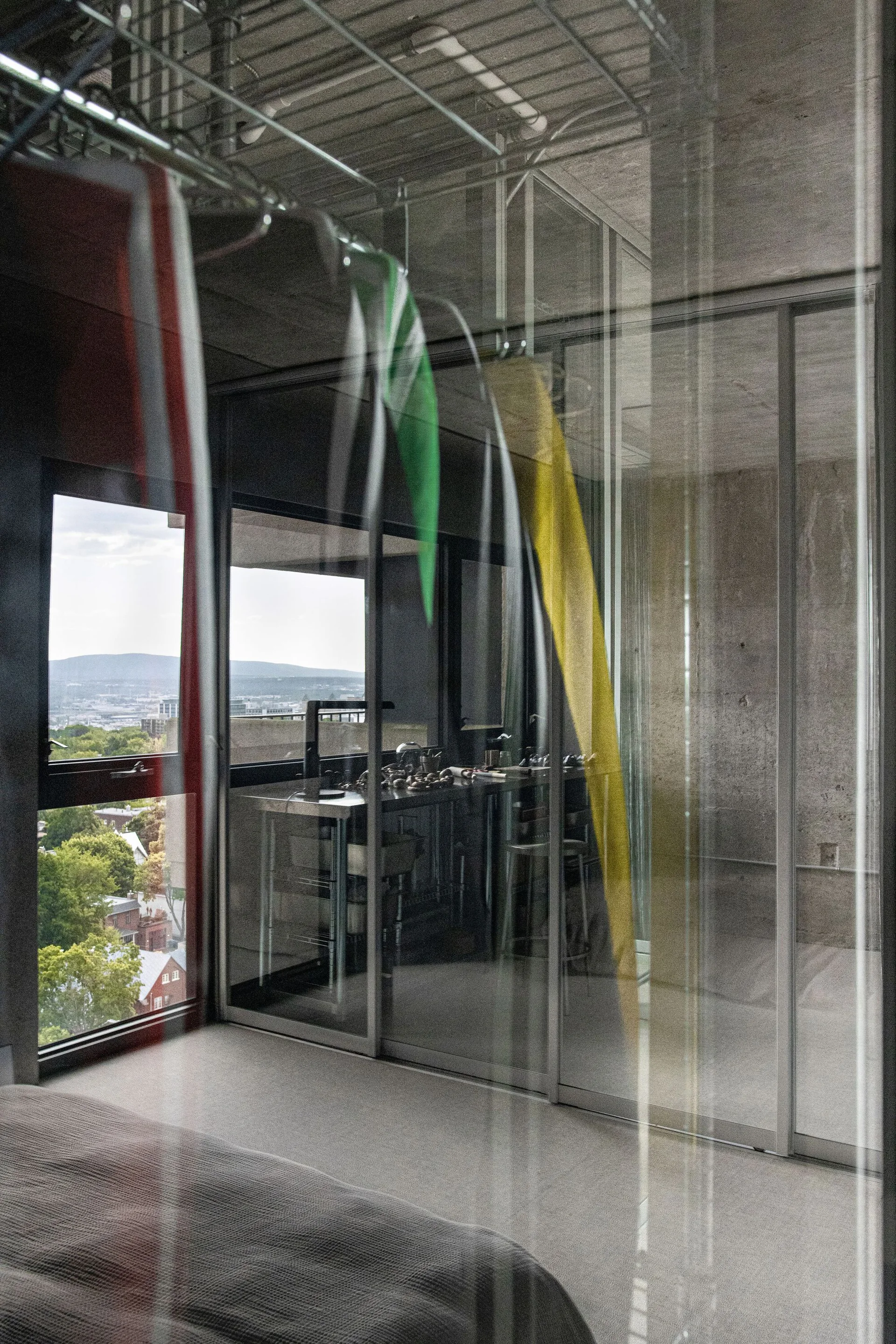 A modern bedroom with a grey bed is separated from an industrial kitchen by a large floor-to-ceiling sliding glass partition wall.