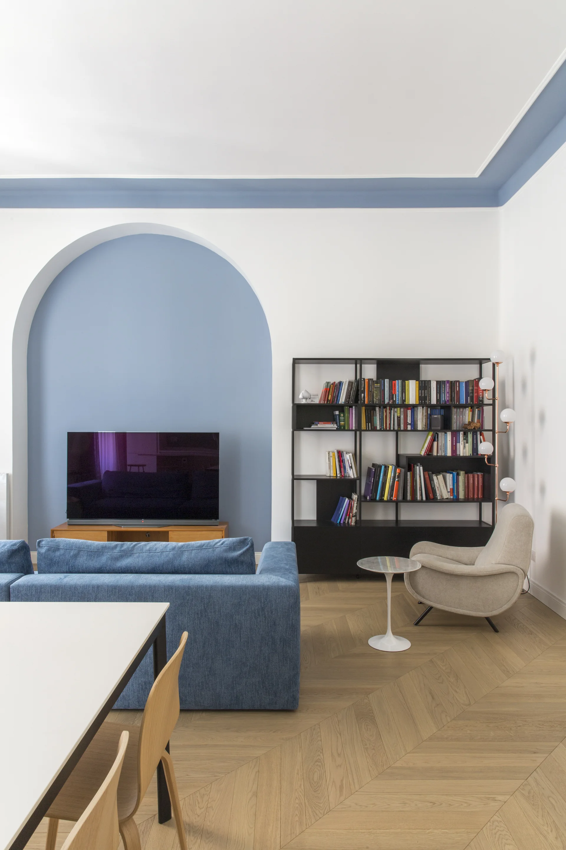 A modern living room featuring a blue sofa, a black metal bookcase, and a television set inside a blue painted wall arch over herringbone wood flooring.