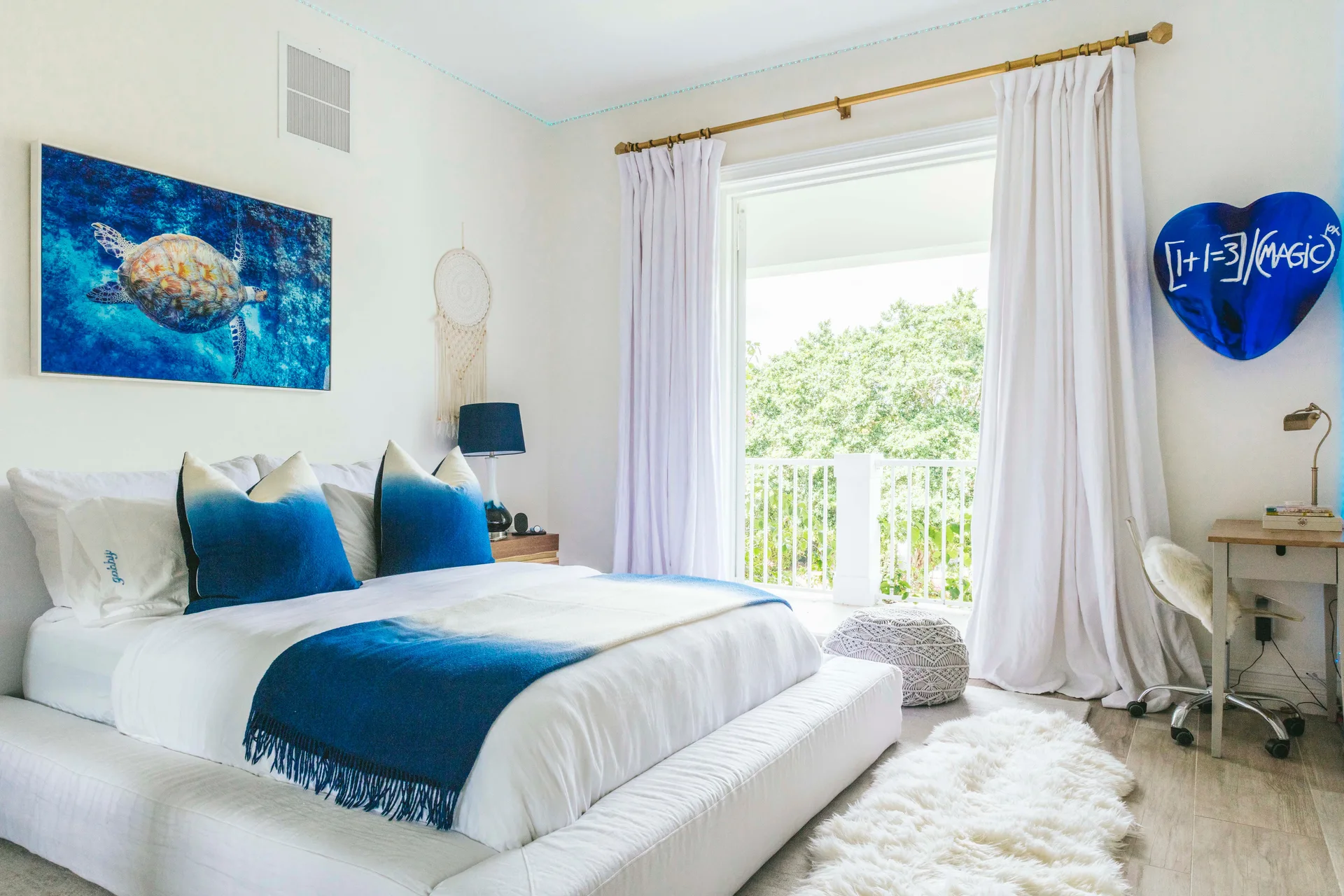A bright white bedroom features a low-profile bed with blue ombre pillows, a faux fur rug, and a large window looking out onto a balcony.