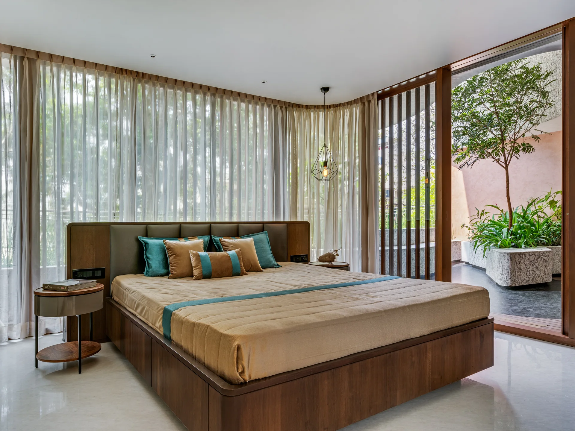 A wooden platform bed with teal and tan bedding is positioned in front of curved sheer curtains overlooking a terrace.