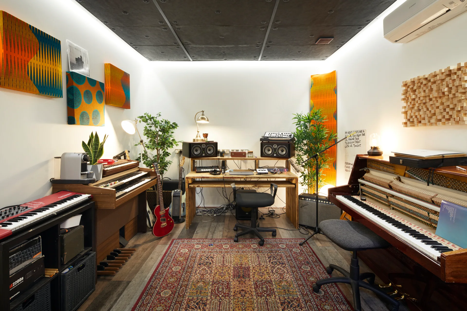 A home music studio featuring a wooden desk, an upright piano with exposed keys, a vintage organ, and colorful acoustic panels on the walls.