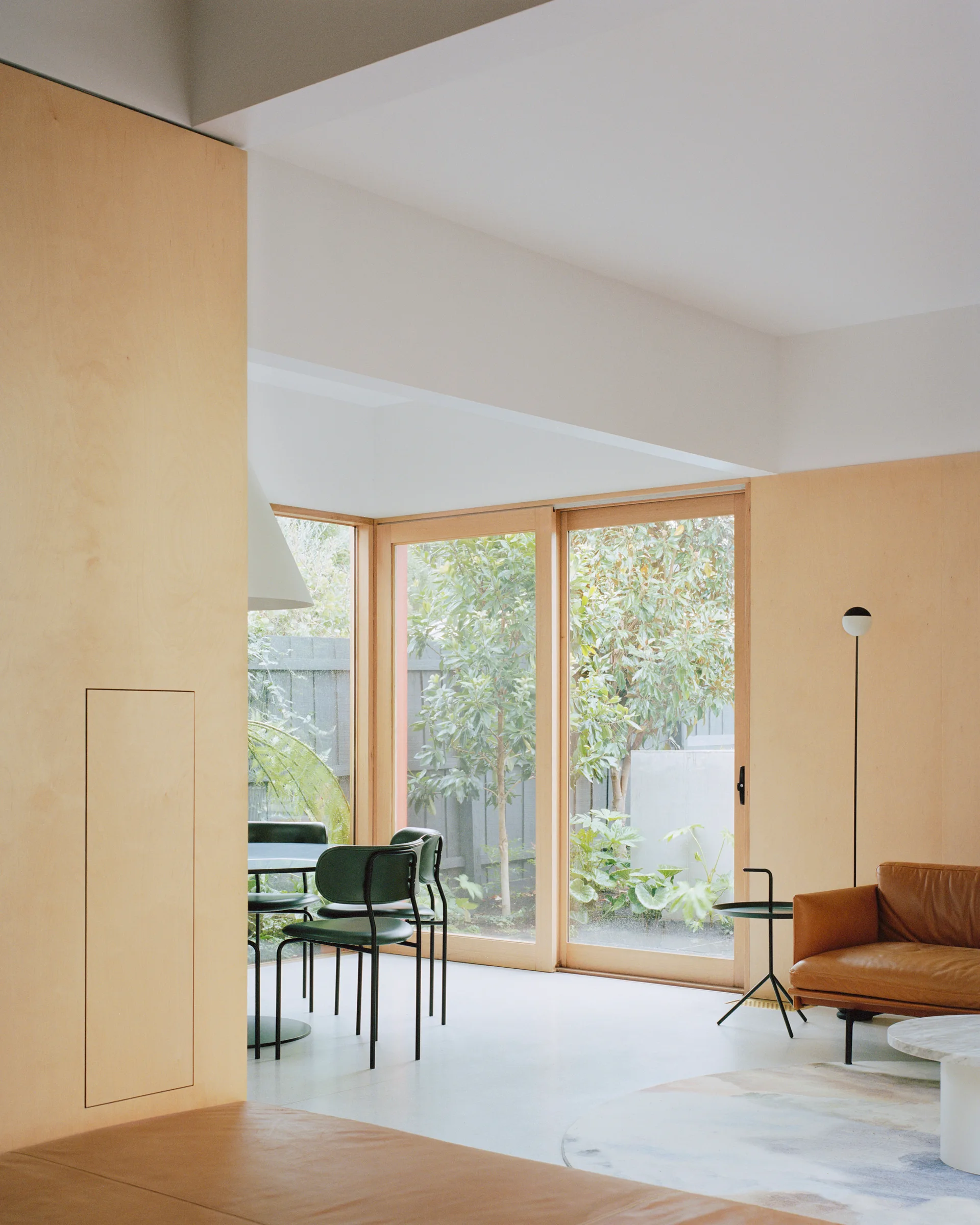 A minimalist open-plan living room featuring light plywood wall panels, a brown leather sofa, and green dining chairs near large glass doors.