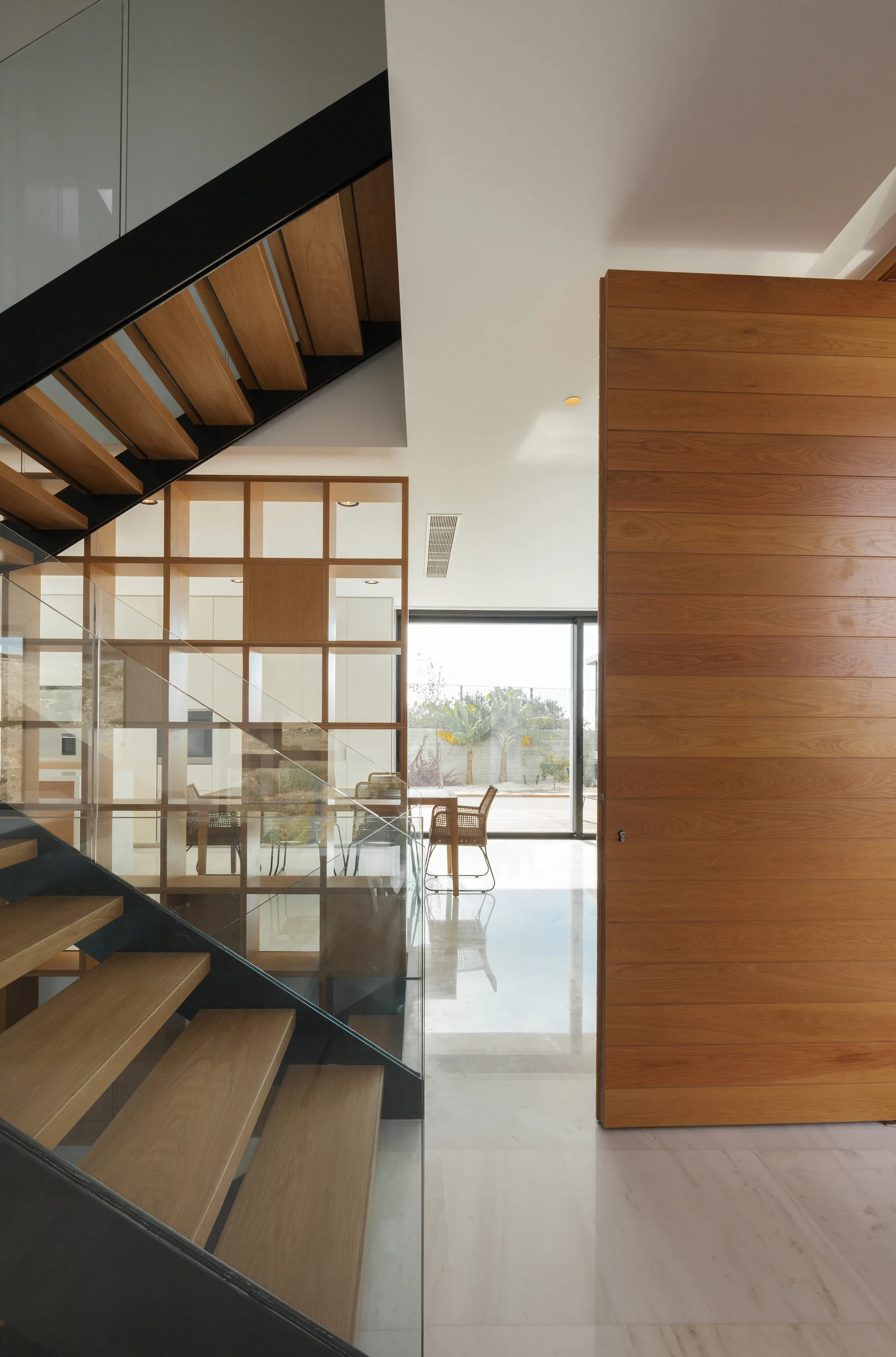 A modern foyer displays a large wooden pivot door, a glass-walled staircase with wood treads, and an open wooden grid room divider in the background.