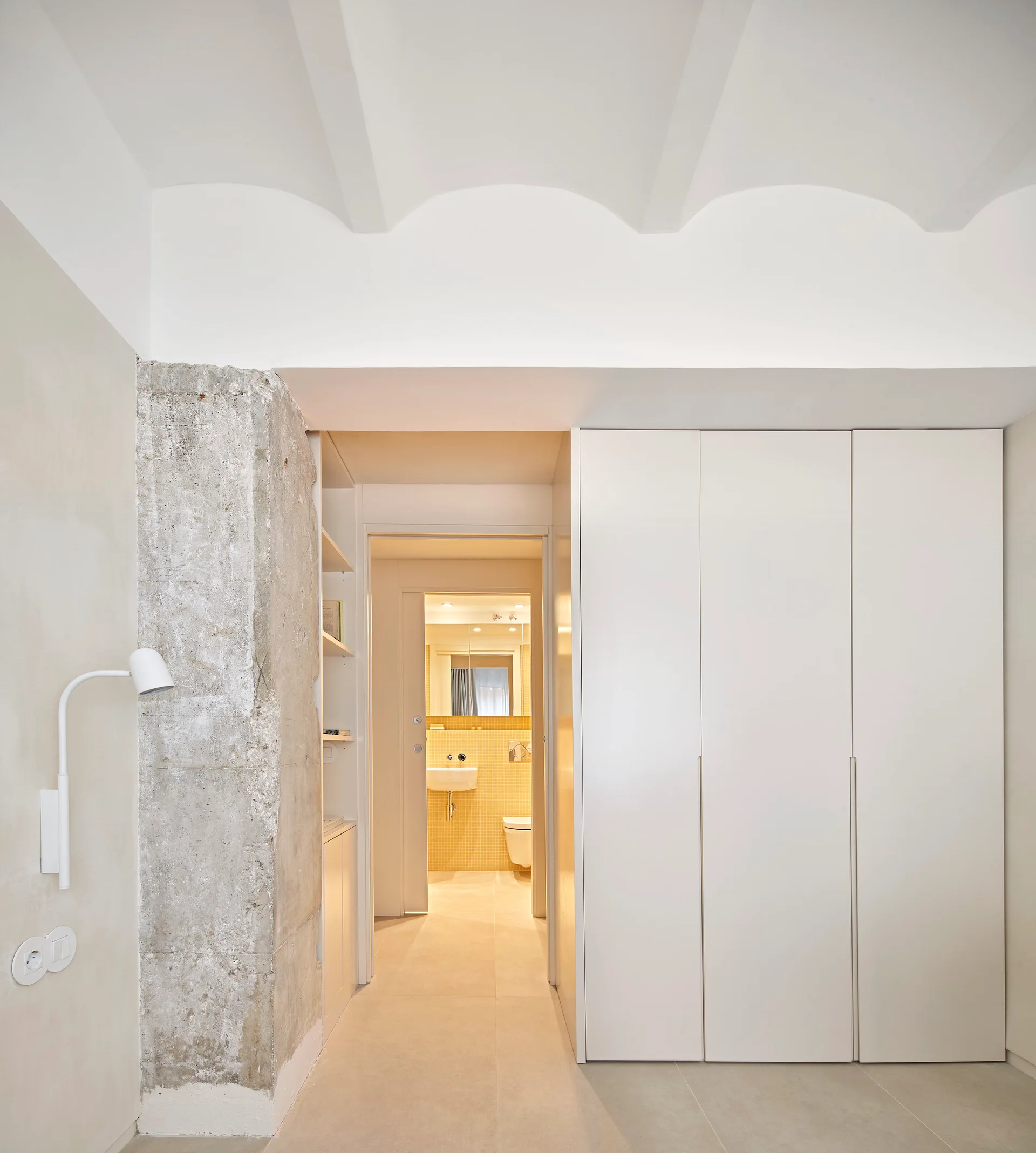 A minimalist interior space features a white built-in wardrobe, an exposed concrete column, and a doorway looking into a yellow-tiled bathroom.