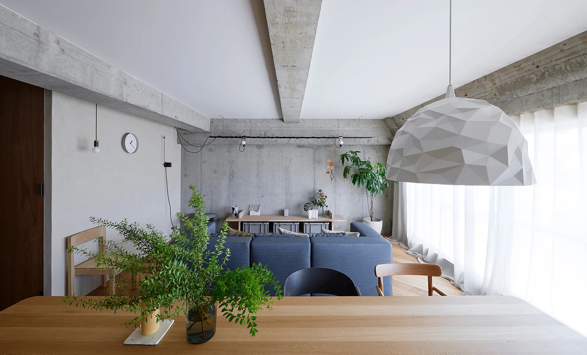 A modern residential living area features an exposed concrete ceiling with beams, a blue sofa, and a large white geometric pendant light.
