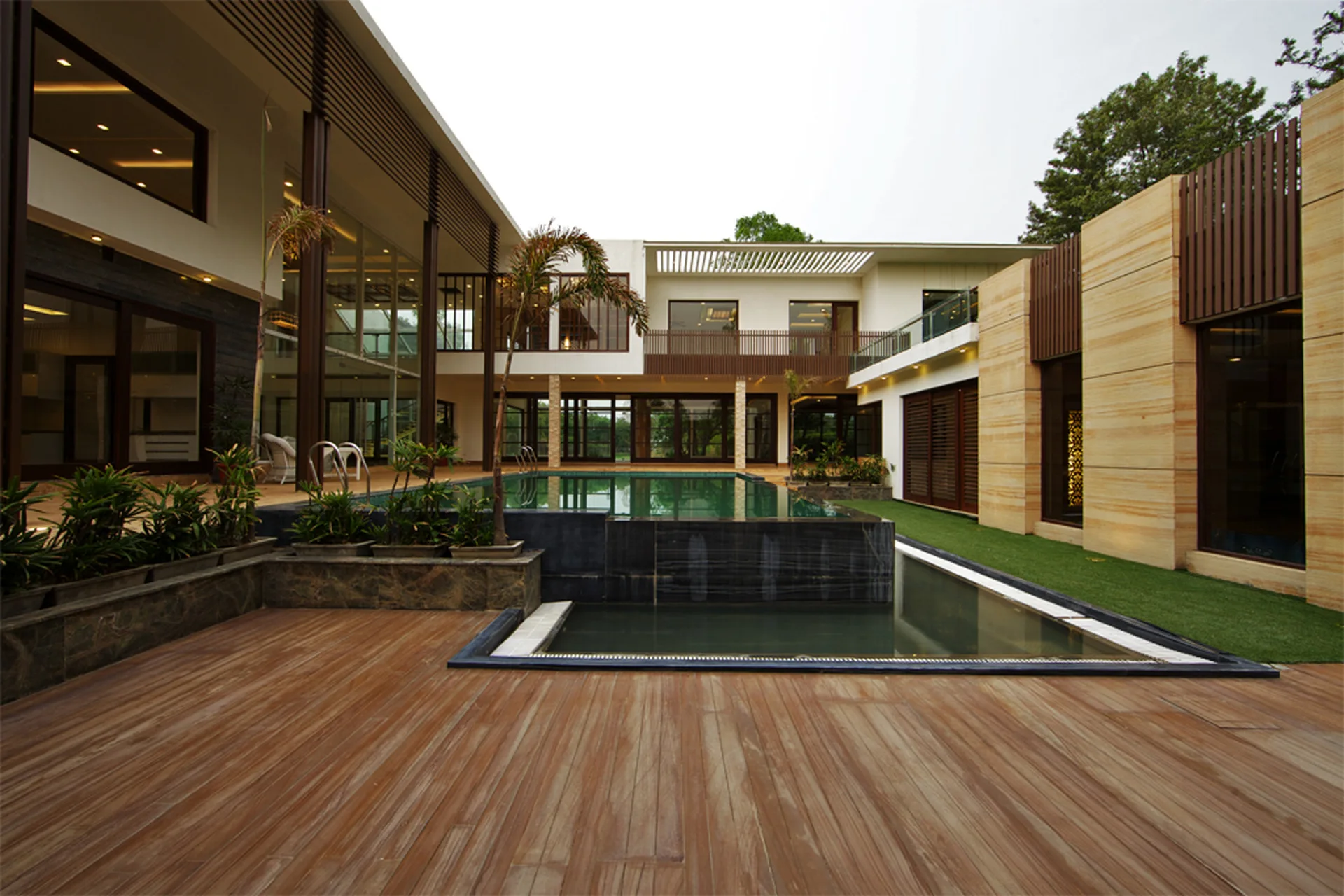 A modern courtyard features a rectangular swimming pool next to a wide wooden deck, surrounded by a two-story residential building with stone walls.
