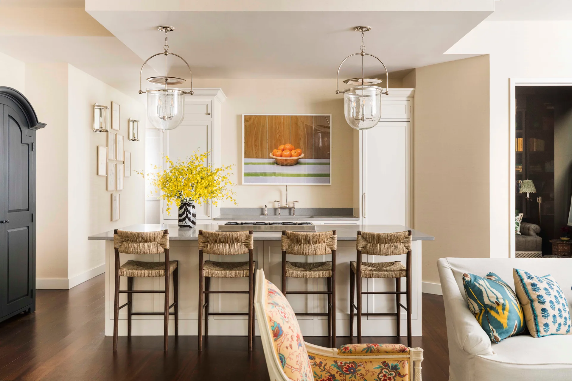 Four woven bar stools line a white kitchen island under two glass lantern pendant lights, set against cream walls and dark wood floors.