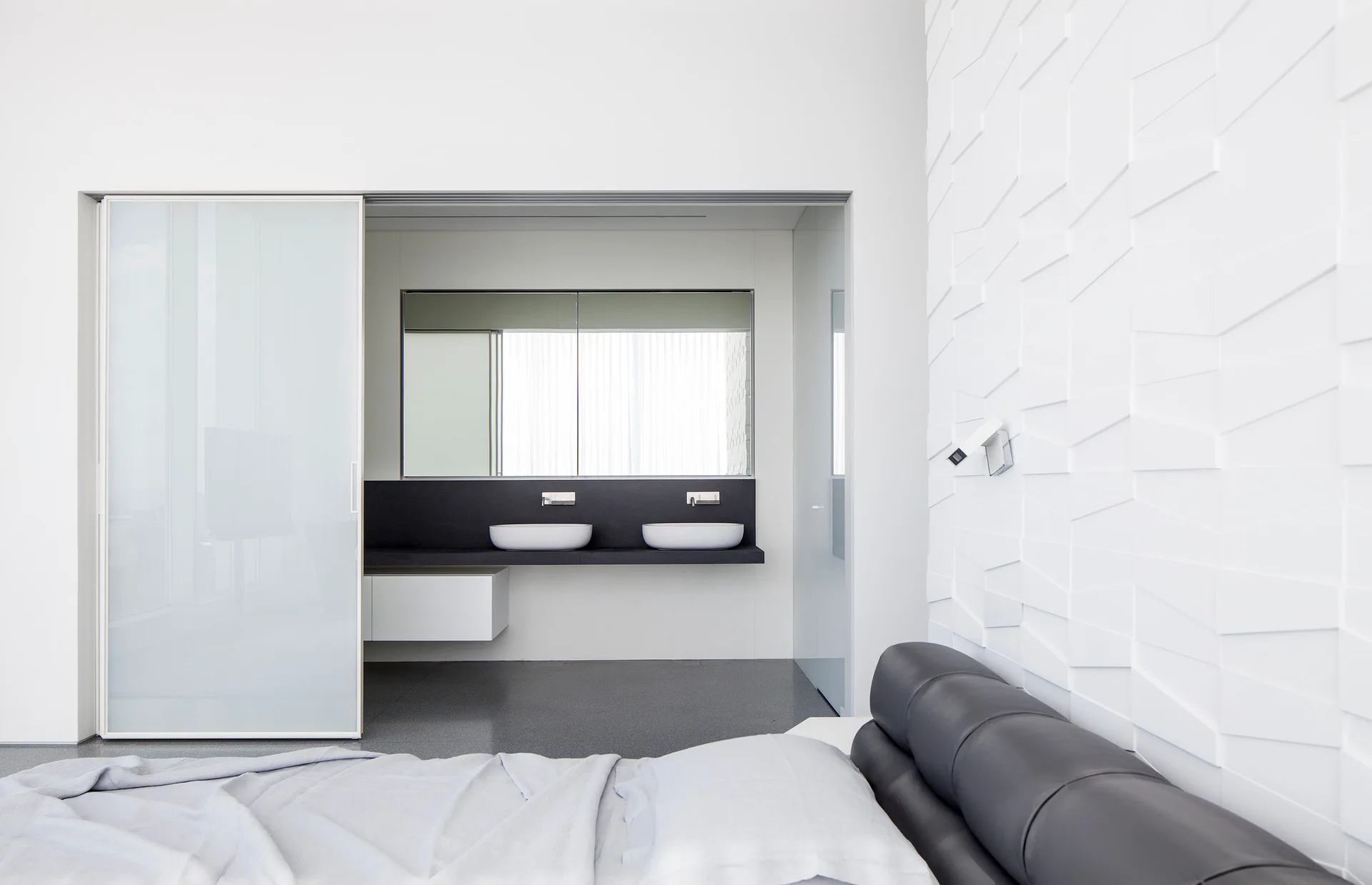 A modern white bedroom featuring a 3D textured accent wall, dark headboard, and an adjacent bathroom with a black double vanity.