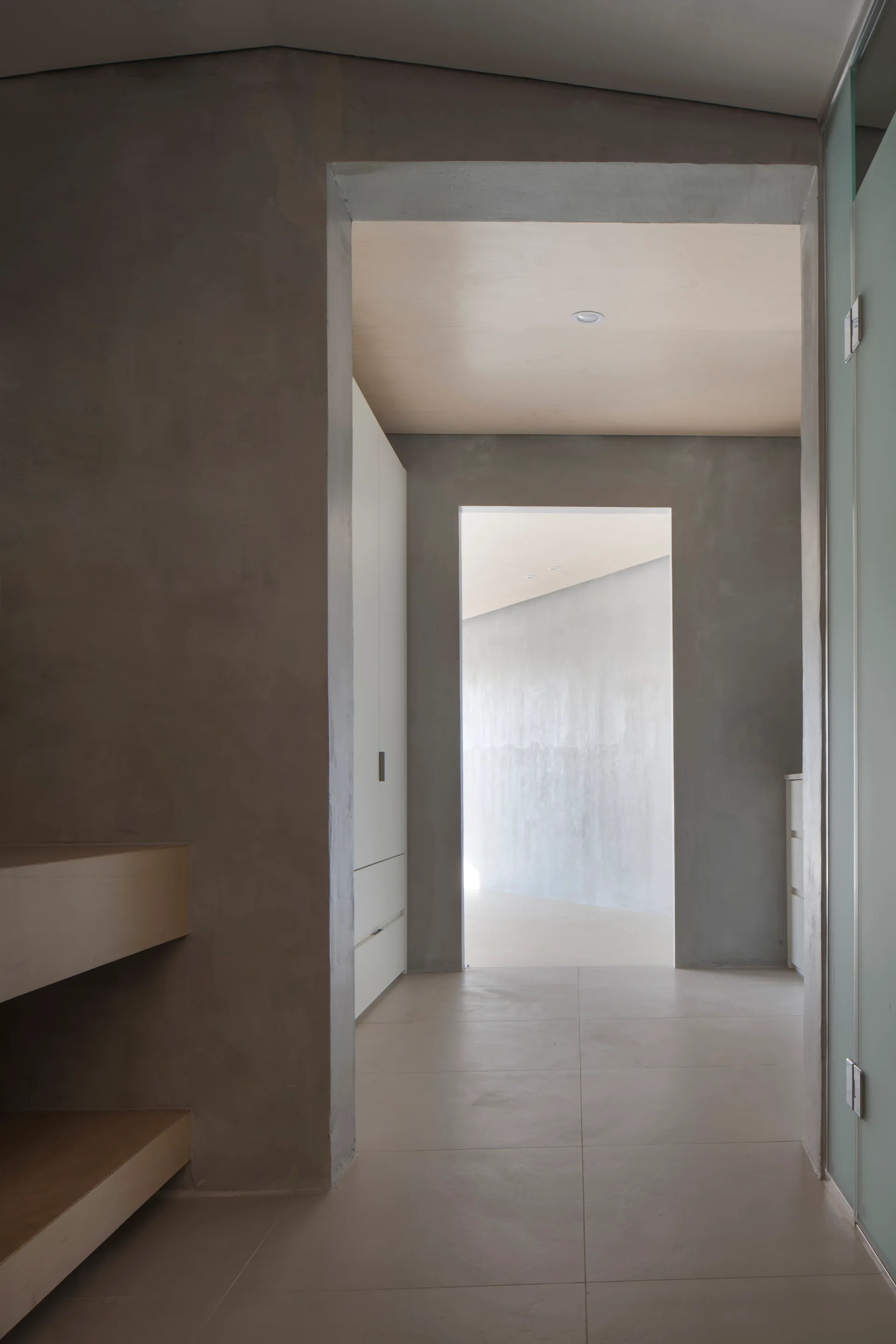 A minimalist hallway features textured gray concrete walls, neutral tile flooring, light wood ceilings, and white built-in cabinetry.