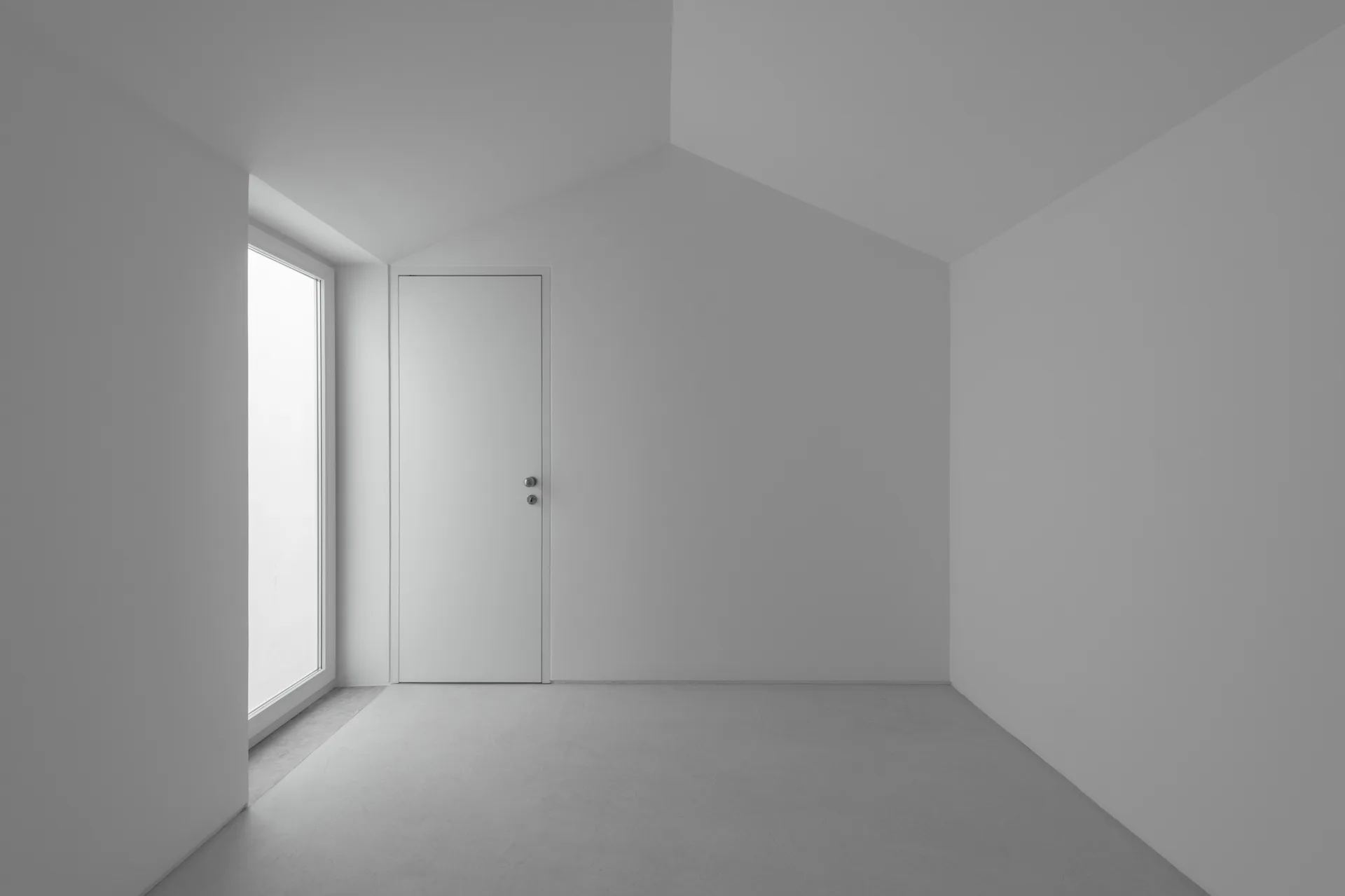 An empty white minimalist room features a sloped ceiling, a flush interior door, and a tall window with natural light.