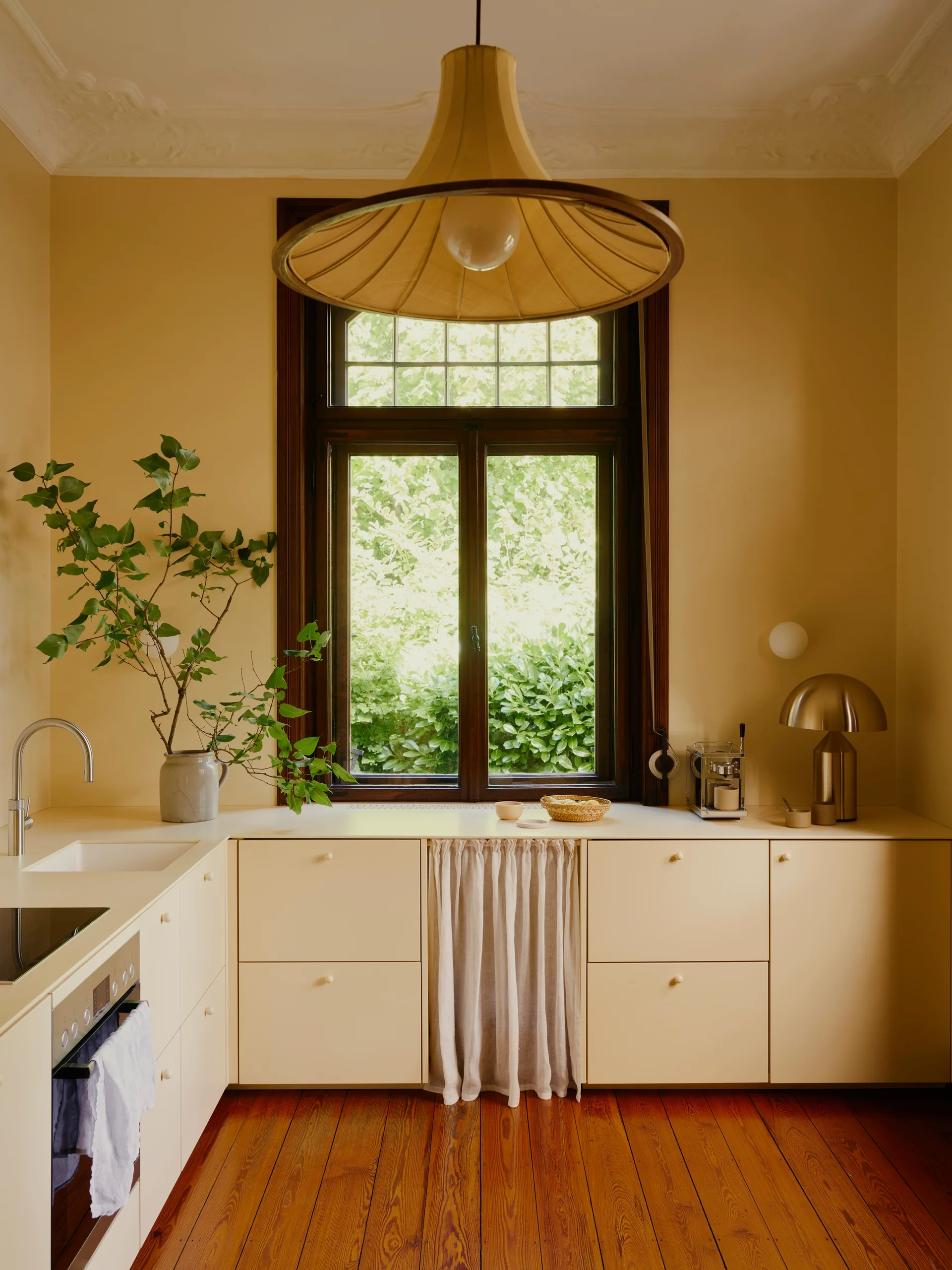 A warm yellow kitchen featuring flat-front base cabinets, a central dark wood window, and a light linen curtain under the countertop.