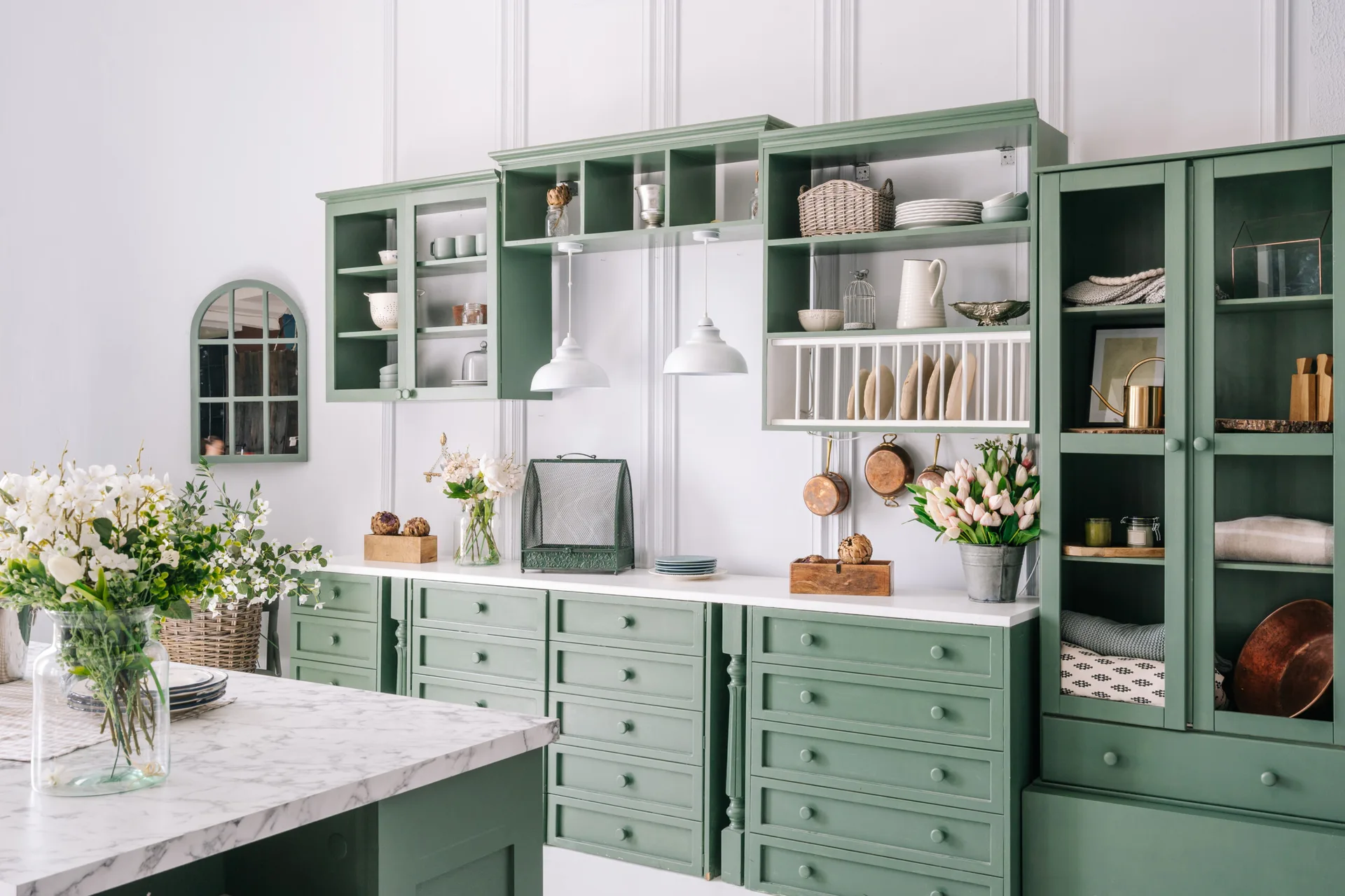 Sage green kitchen cabinetry with a built-in plate rack, white marble-topped island, white pendant lights, and hanging copper cookware.