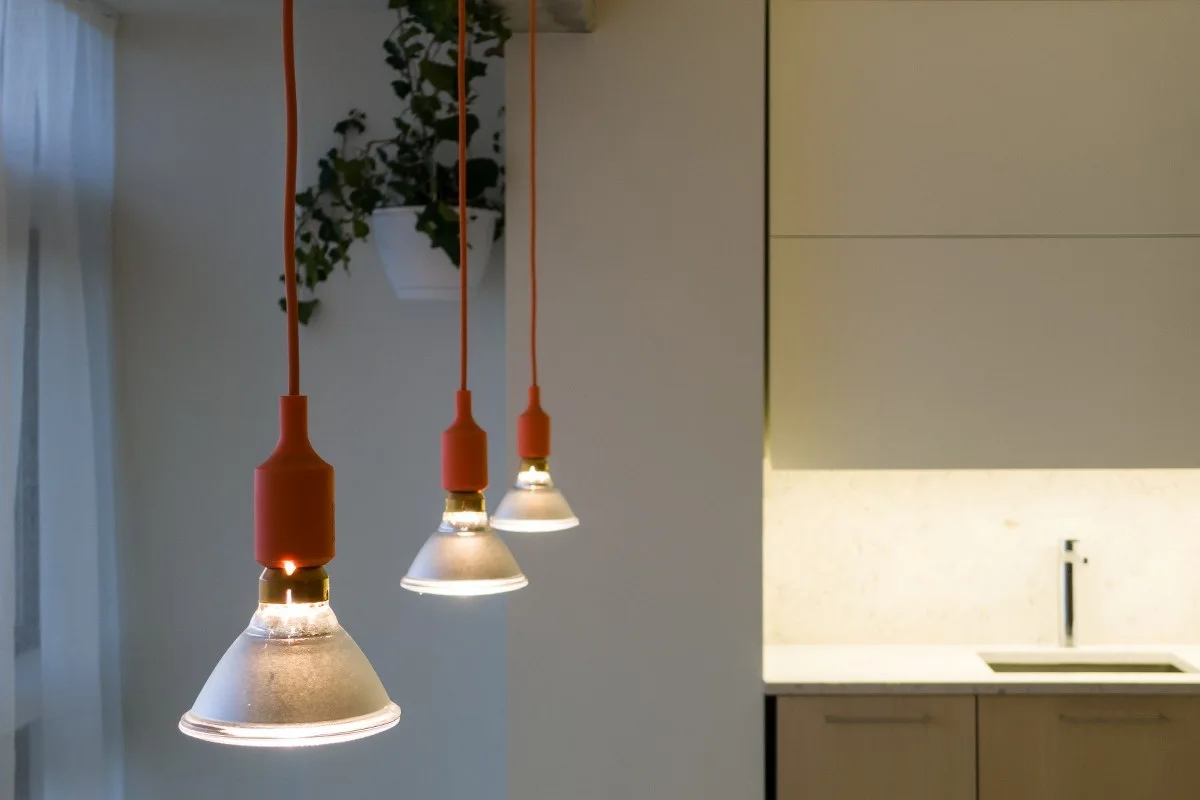 Three pendant lights with orange cords hang in front of a modern kitchen featuring beige cabinets, under-cabinet lighting, and a silver faucet.