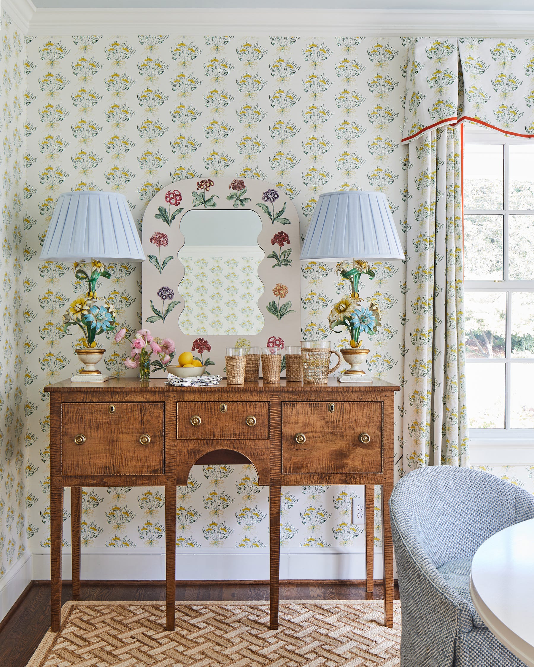 A wooden console table stands against yellow floral wallpaper beneath a floral-bordered scalloped mirror and two lamps with blue pleated shades.