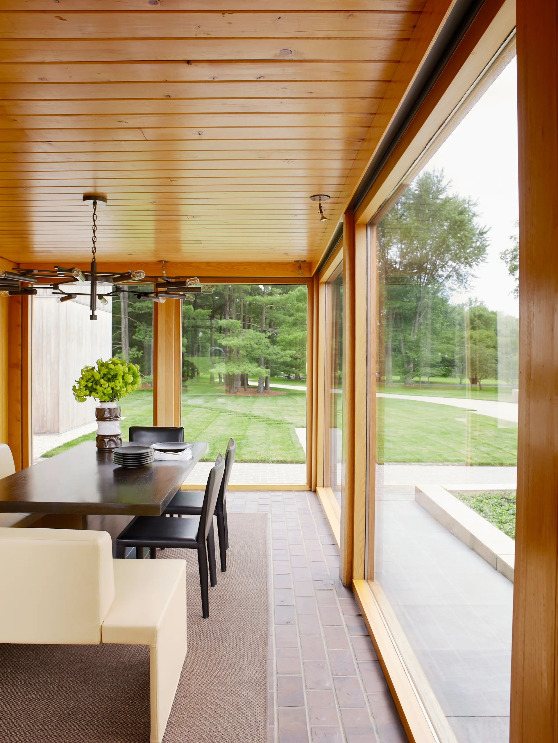 A modern dining room features a dark wood table with black chairs, a wood-paneled ceiling, and floor-to-ceiling glass walls looking out at a lawn.