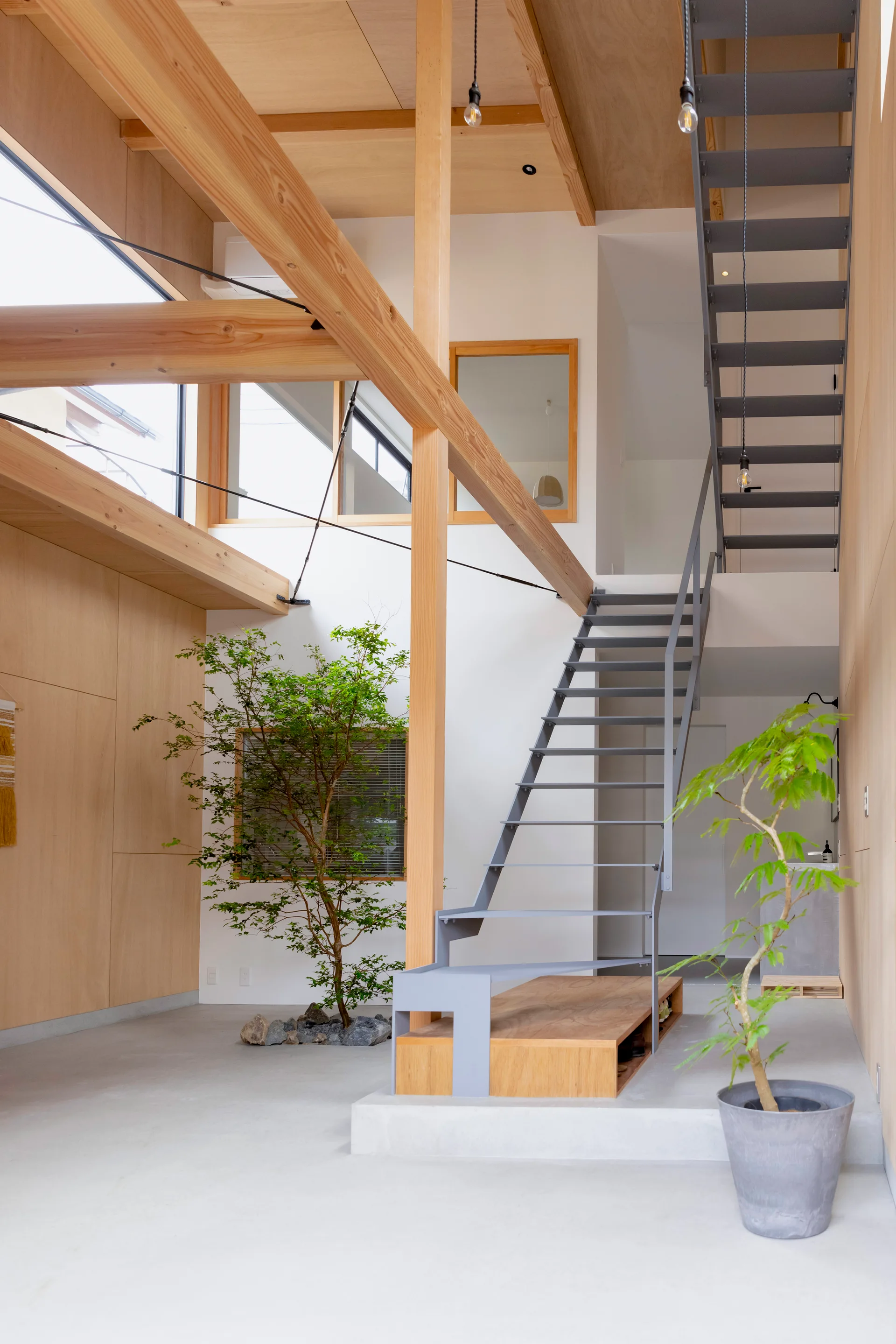 A modern home interior features an open-tread steel staircase next to an indoor tree planted in a small rock bed under high wood ceilings.
