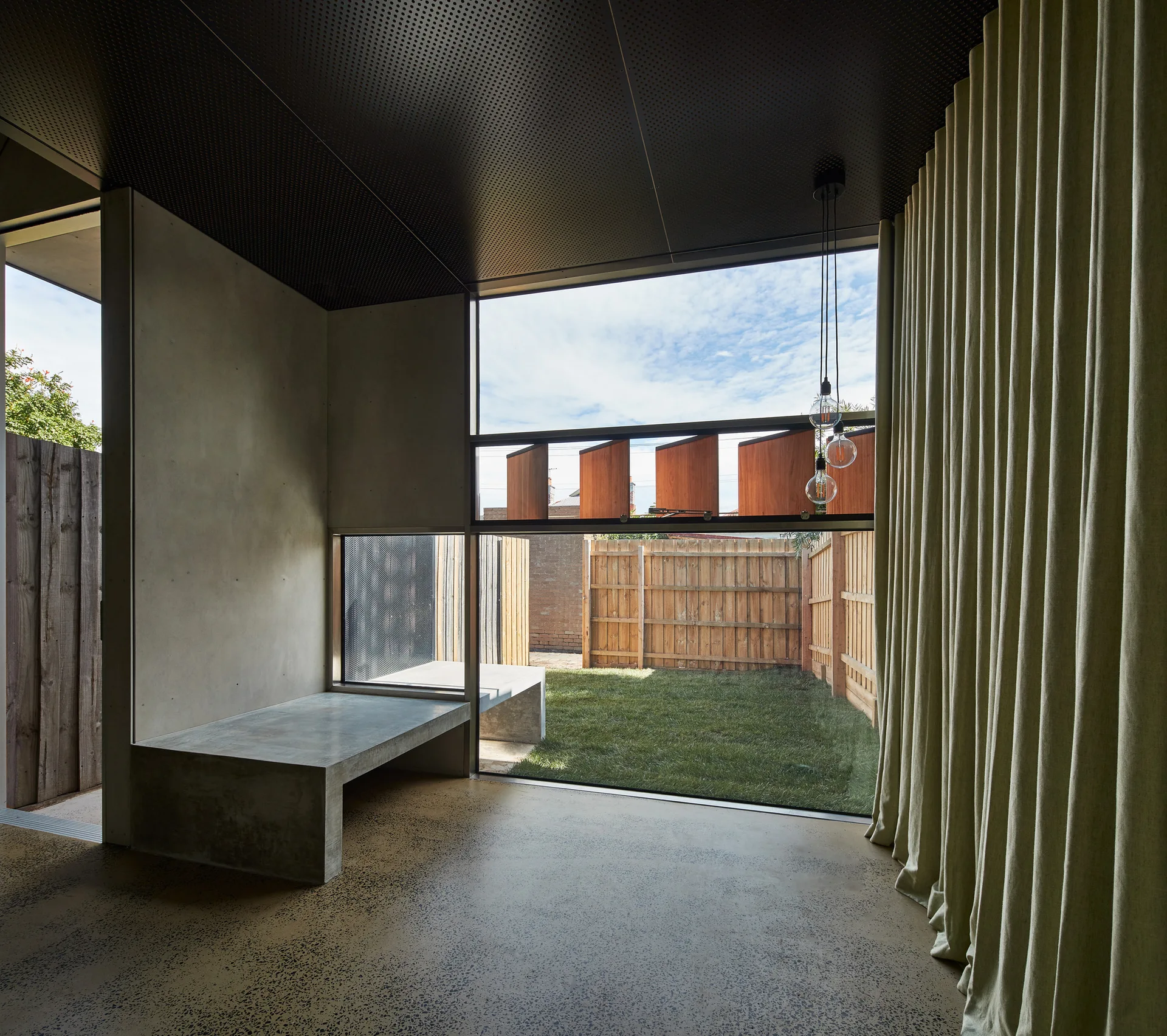 A minimalist room features concrete walls, a built-in concrete bench, a dark perforated ceiling, and large glass windows looking out to a grassy yard.