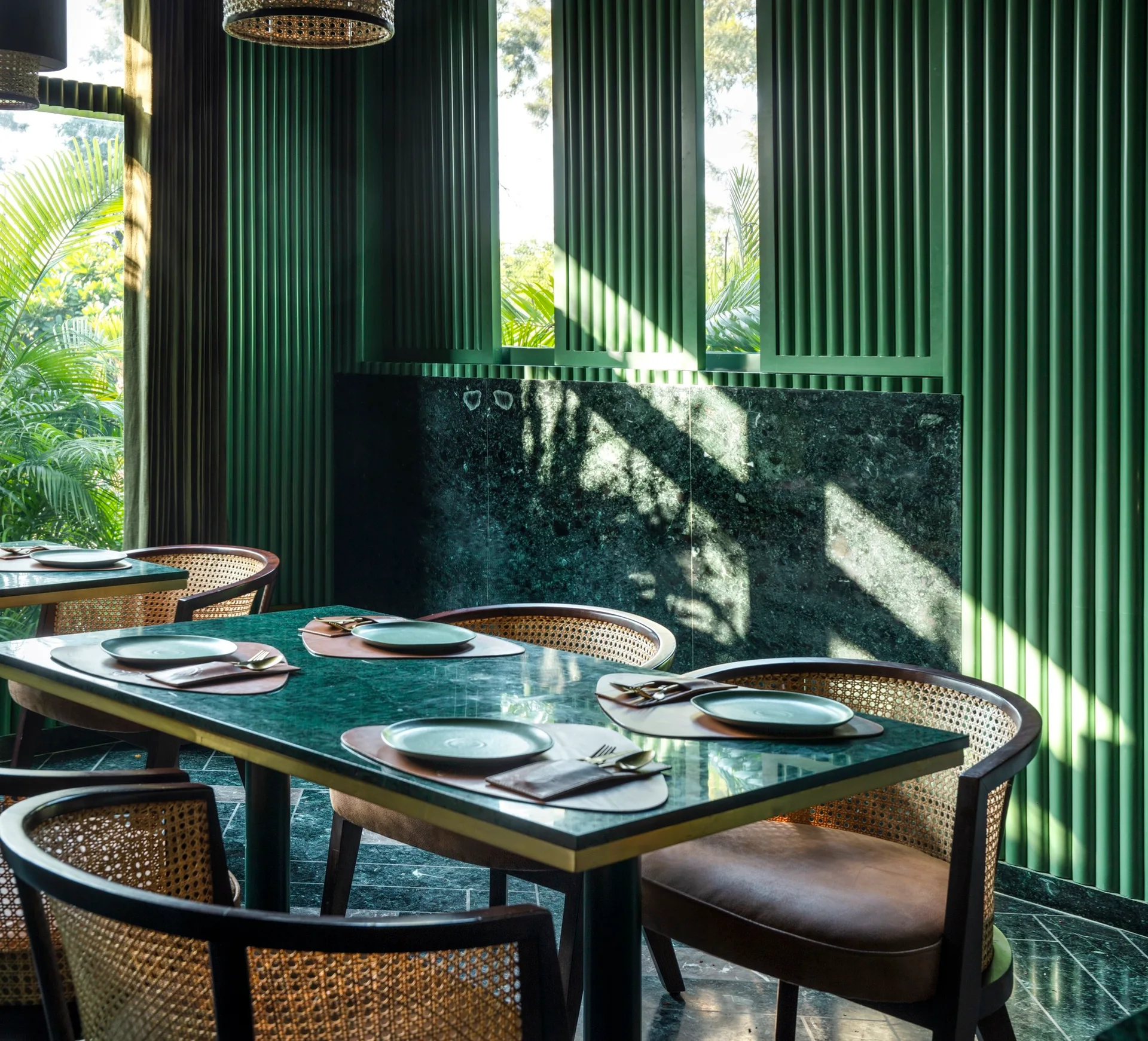 A dining room features green marble-topped tables set with plates, surrounded by wicker and leather chairs against green slatted wood paneled walls.