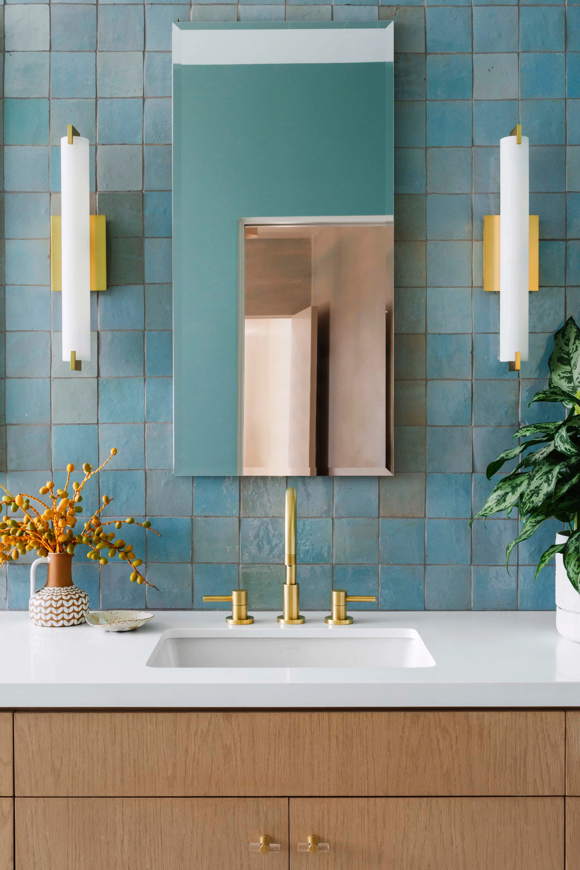 A modern bathroom vanity featuring a warm wood cabinet, white countertop, brass faucet, rectangular mirror, and a wall of square blue tiles.