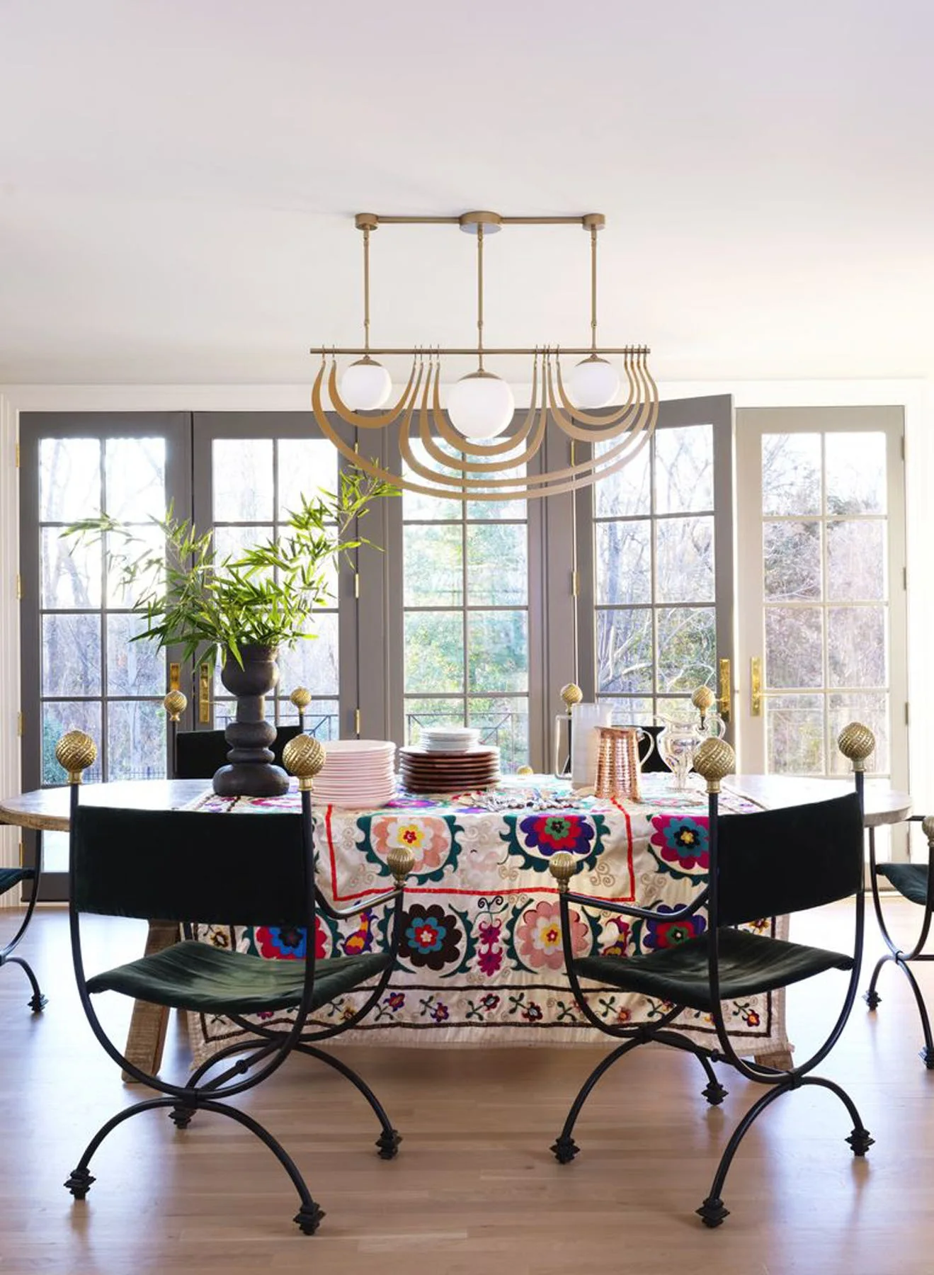 A dining room features black metal chairs around a table with a colorful floral embroidered tablecloth under a modern brass chandelier.