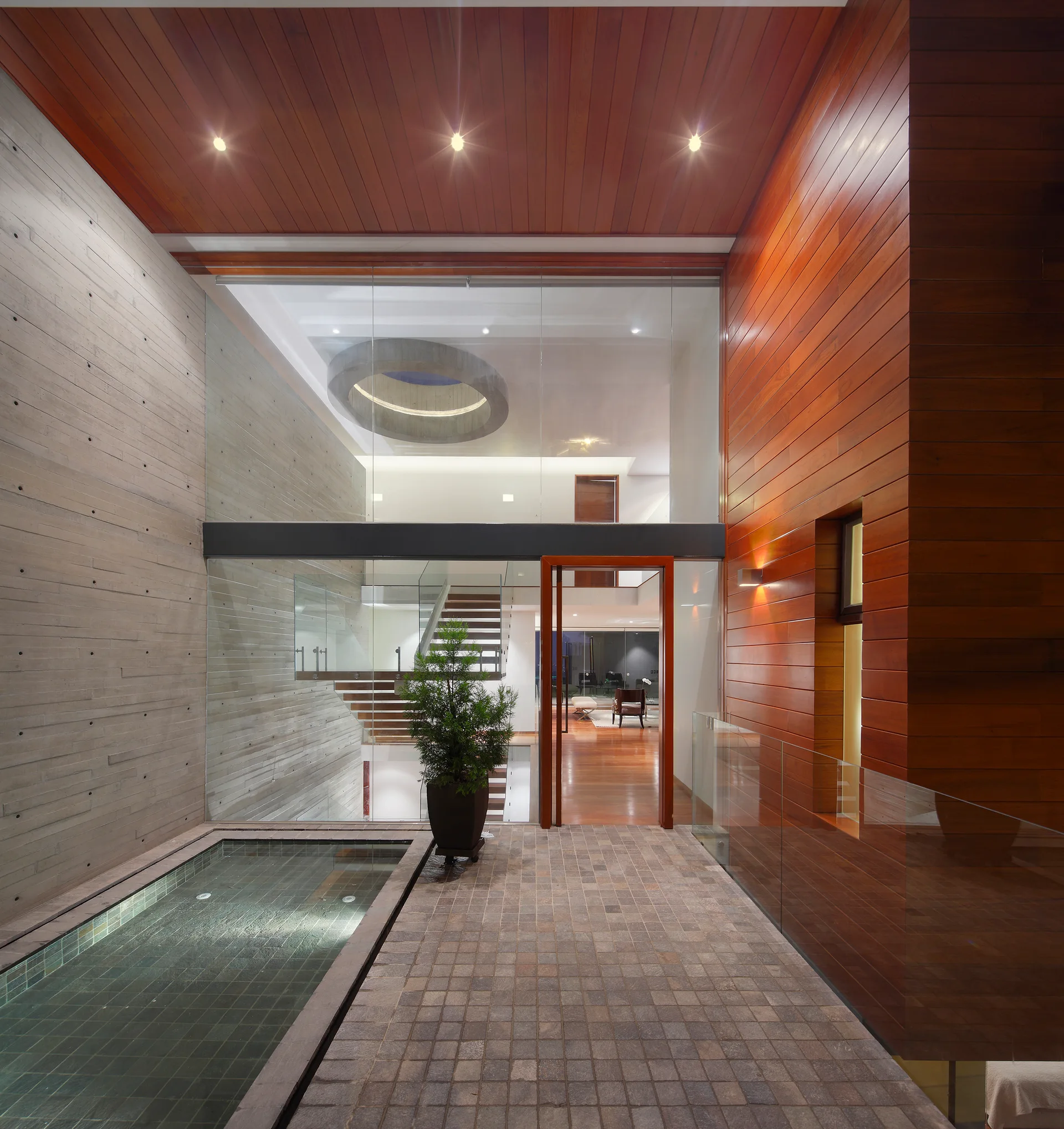 A modern entryway features board-formed concrete walls, warm wood ceiling panels, a tiled stone floor with a water pool, and a large glass partition.