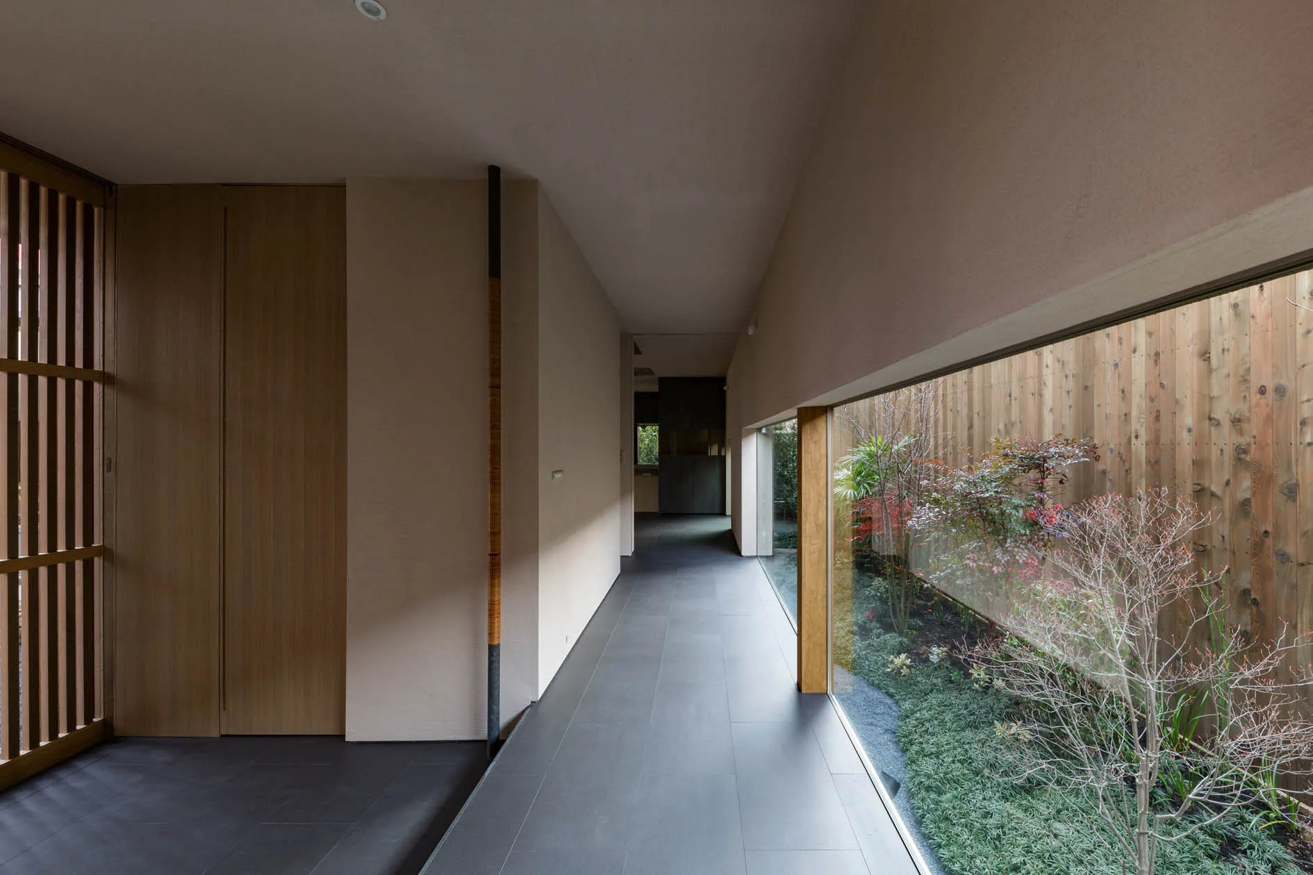 A long minimalist hallway features grey tiled flooring, beige walls, and a large horizontal window looking out onto a narrow garden with a wooden fence.