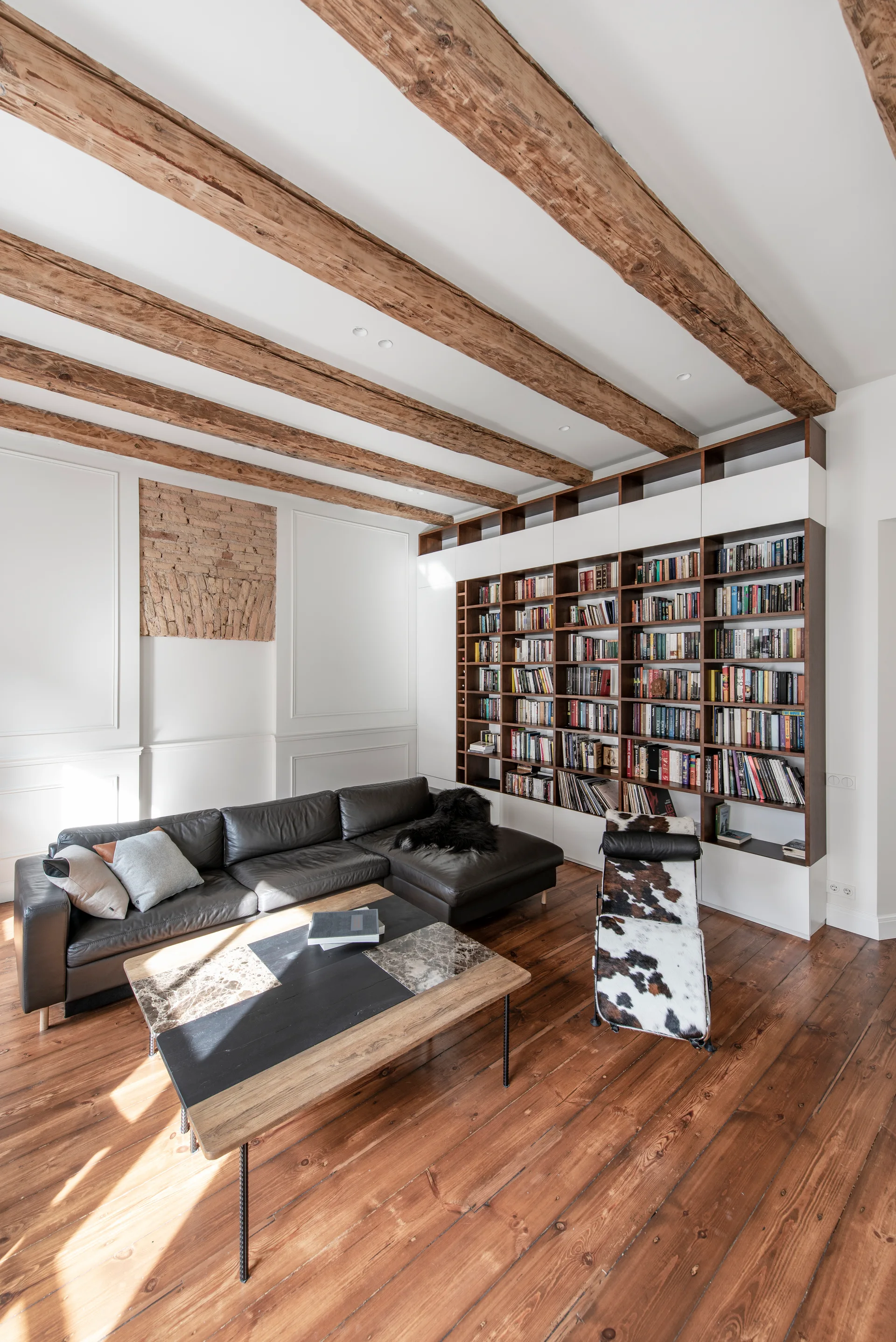 A living room features a dark leather sectional sofa, a massive built-in bookshelf, exposed wooden ceiling beams, and a patterned lounge chair.