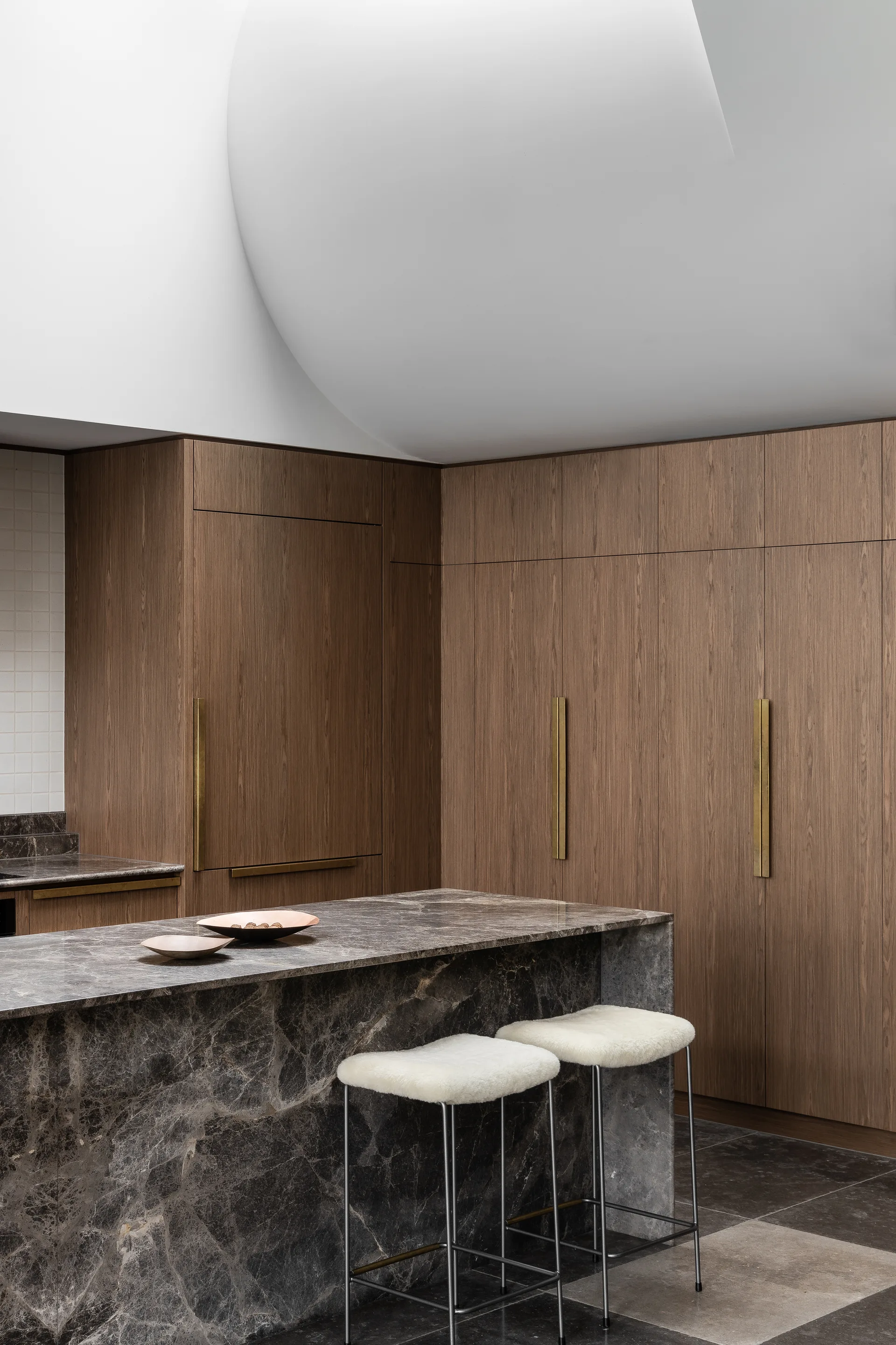 A modern kitchen featuring full-height wood cabinetry with long gold handles, a dark grey veined marble island, and two fuzzy white bar stools.