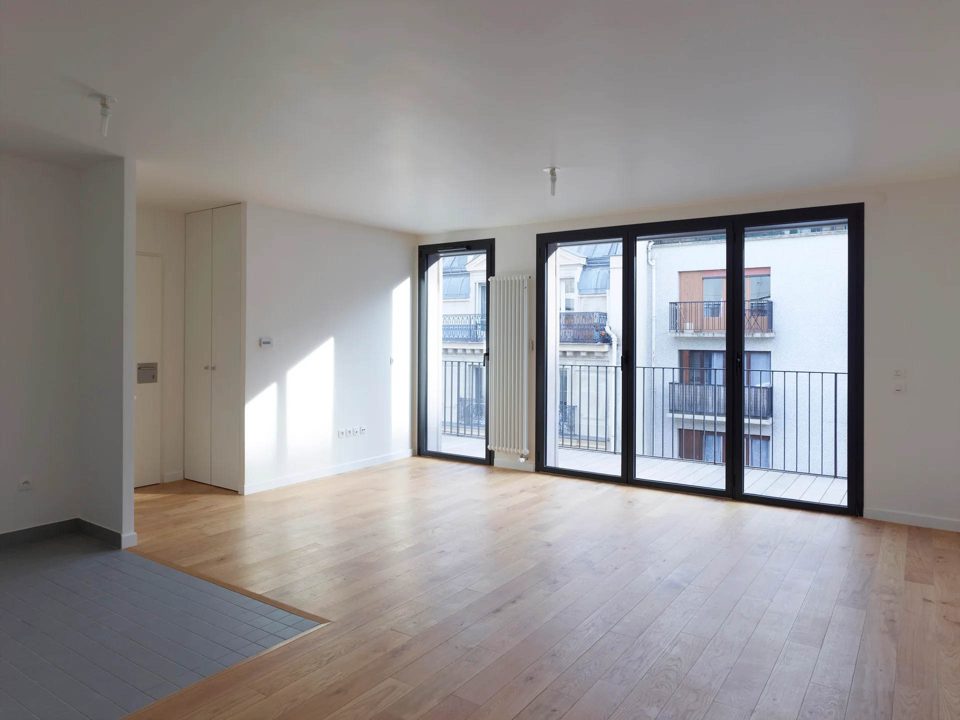 An empty modern apartment room with light wood flooring, white walls, and black-framed balcony glass doors.
