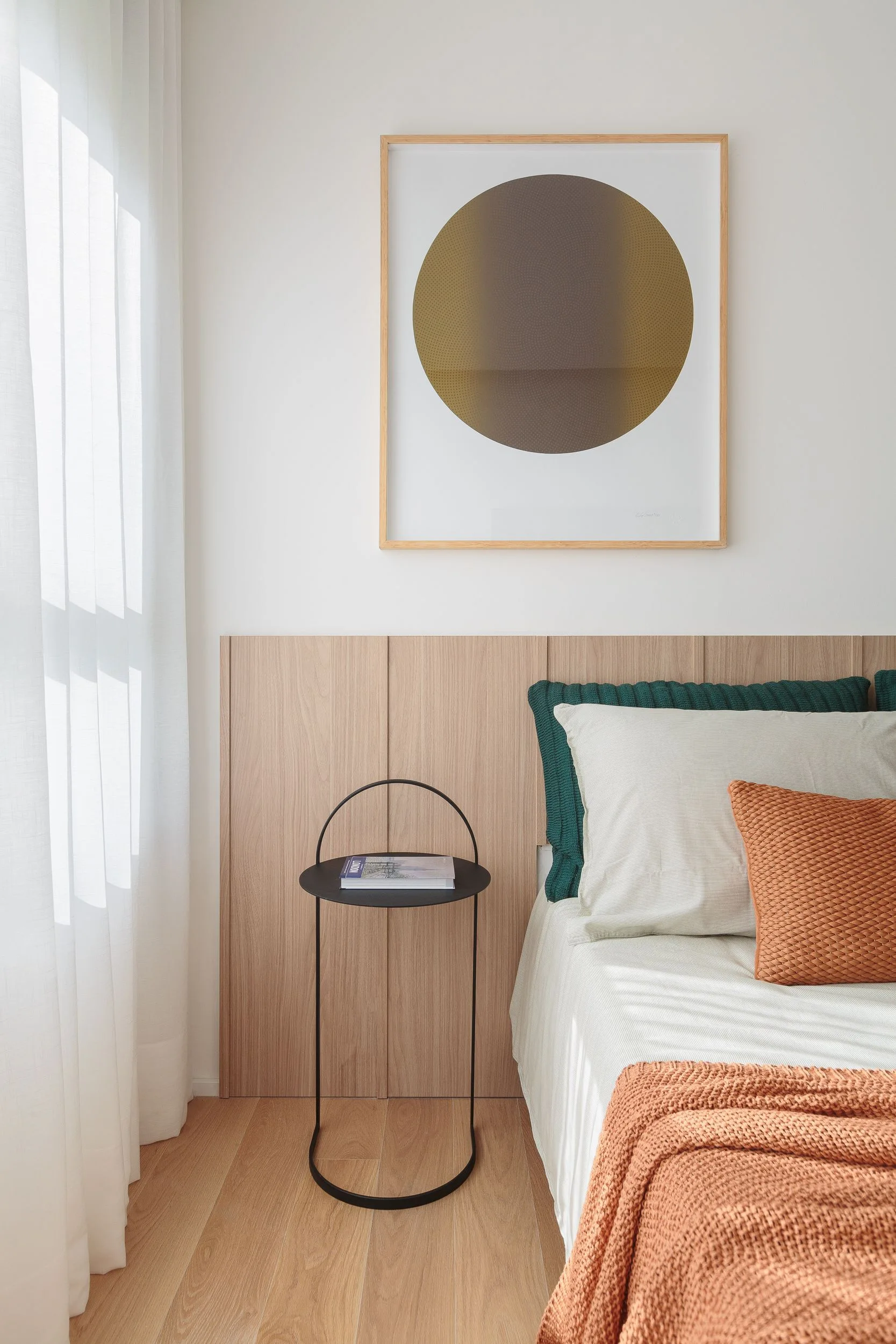 A bedroom featuring a low wood-paneled headboard, a minimalist black metal side table, and textured bedding in earthy orange and green tones.