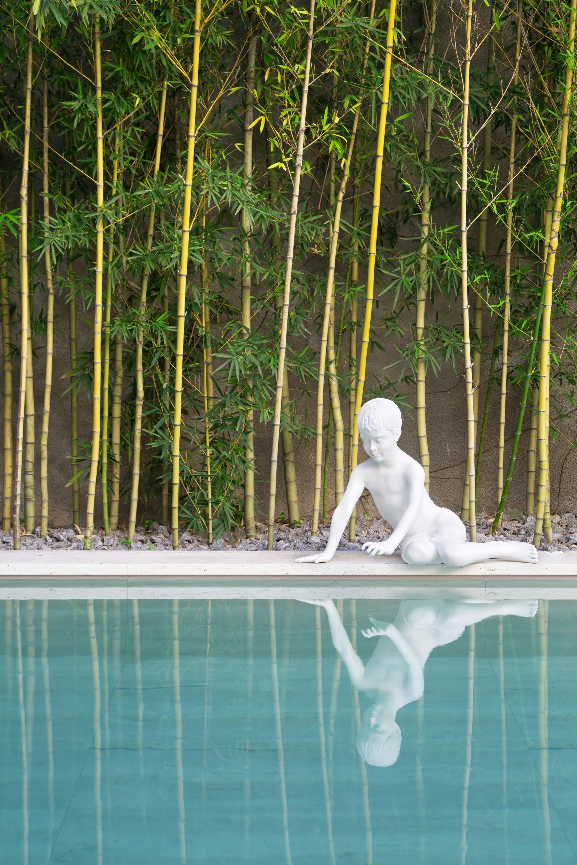 A white sculpture of a boy sits on the stone edge of a swimming pool, reflected in the water against a background of tall green bamboo.