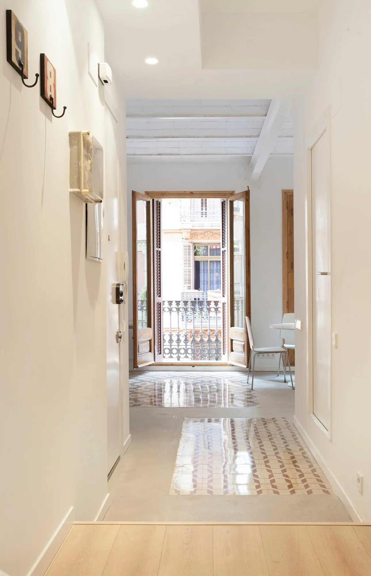 A white hallway with light wood flooring transitions to glossy geometric patterned floor tiles leading to open wooden double doors and a balcony.