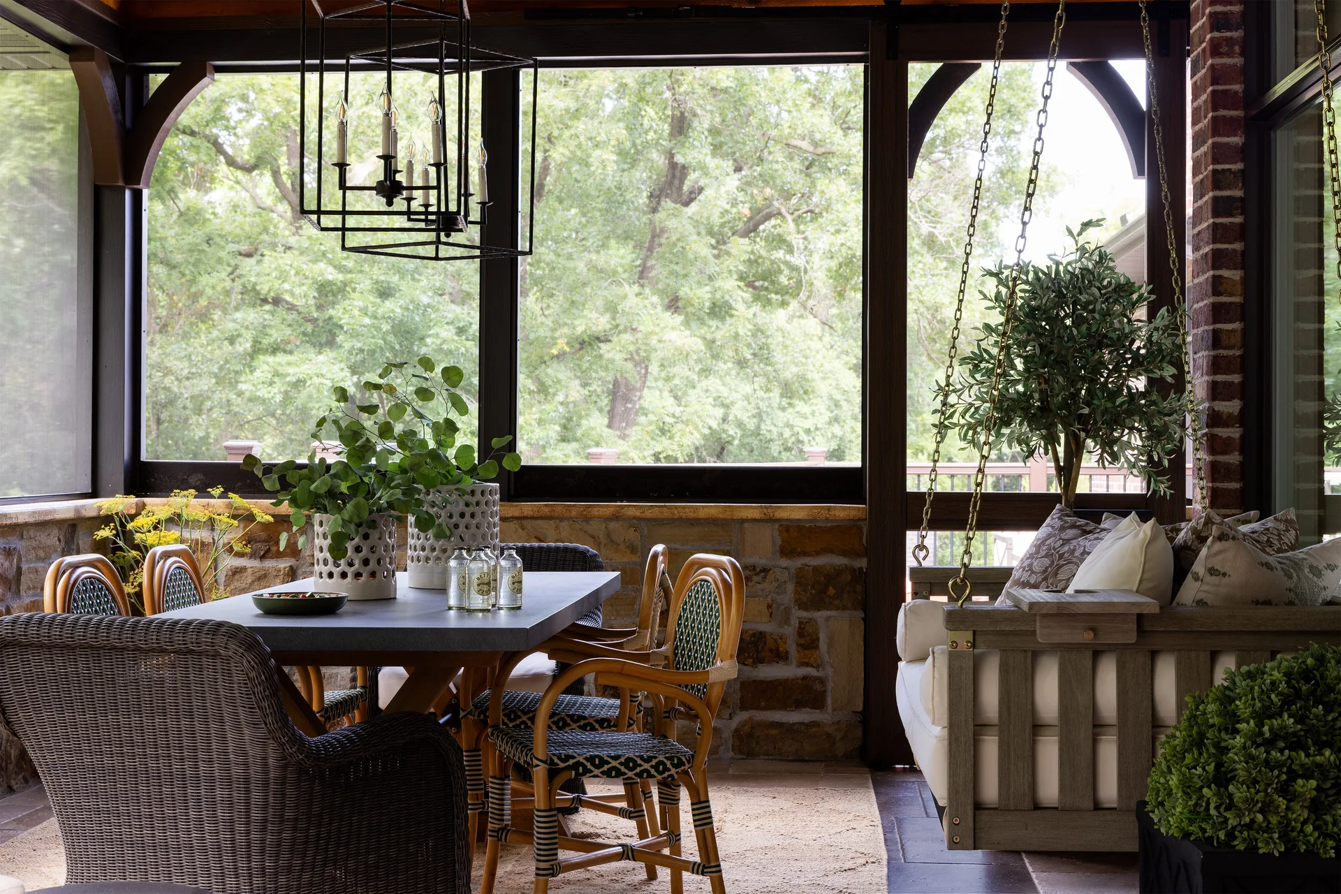 A screened-in porch features a rectangular dining table with wicker chairs next to a hanging wooden porch swing with throw pillows.