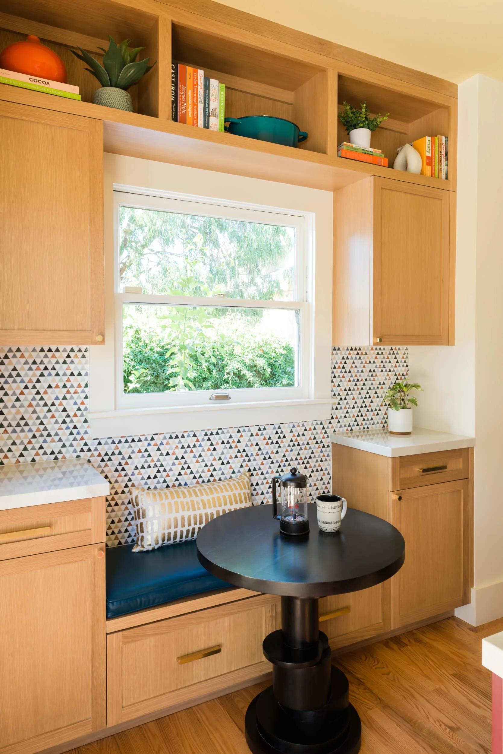 A wooden breakfast nook with built-in cabinetry framing a window, a geometric tiled backsplash, a cushioned bench, and a small black round table.