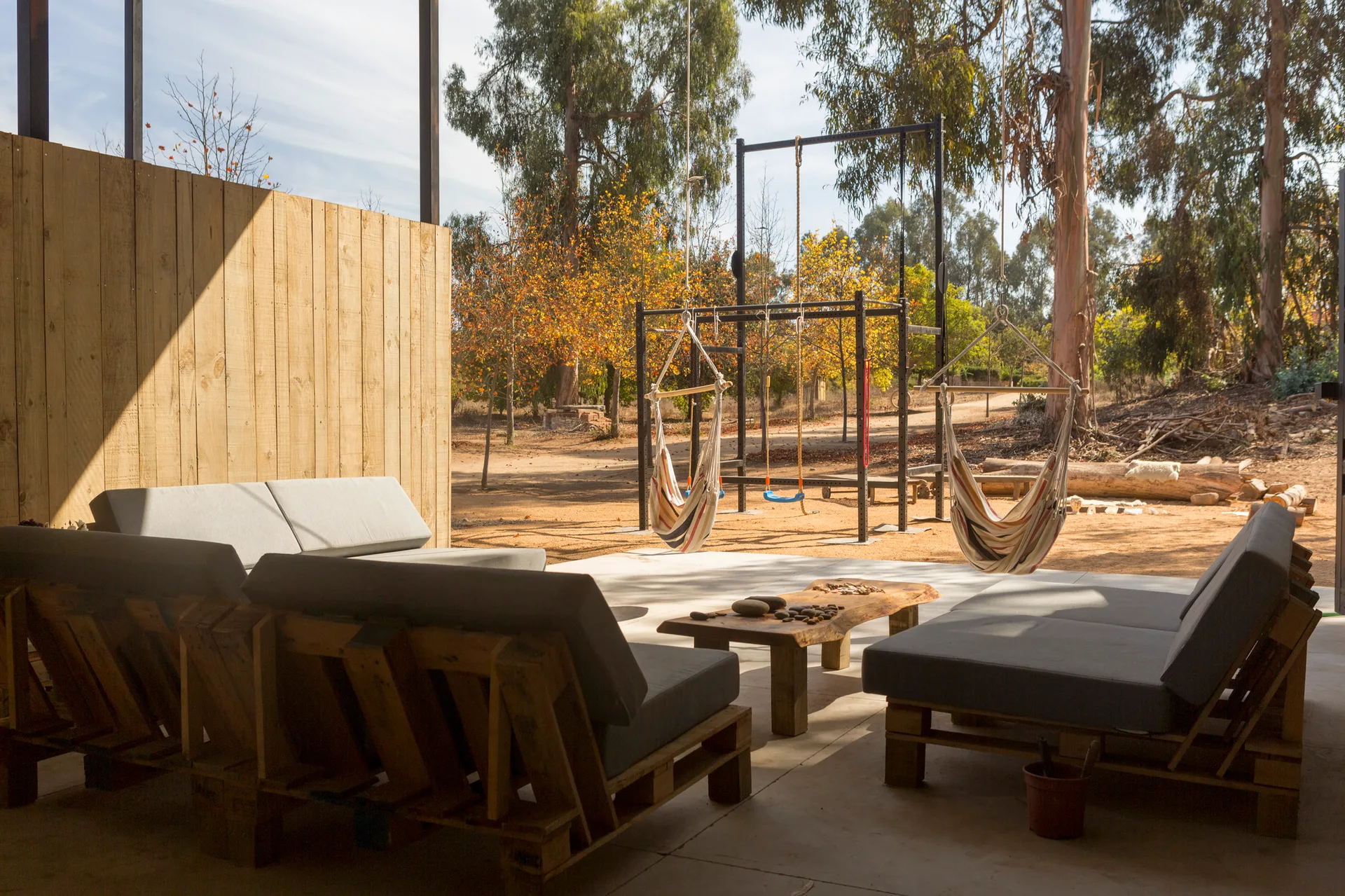 A covered concrete patio features grey pallet sofas, a live-edge wooden coffee table, and striped hammock chairs overlooking a rustic dirt yard.