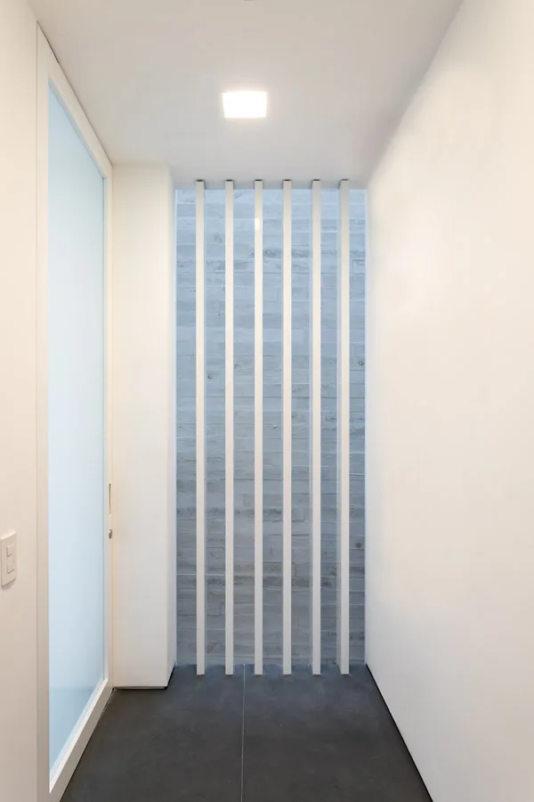 A minimalist hallway features dark stone floor tiles, smooth white walls, a frosted glass door, and vertical white slats in front of a textured grey wall.