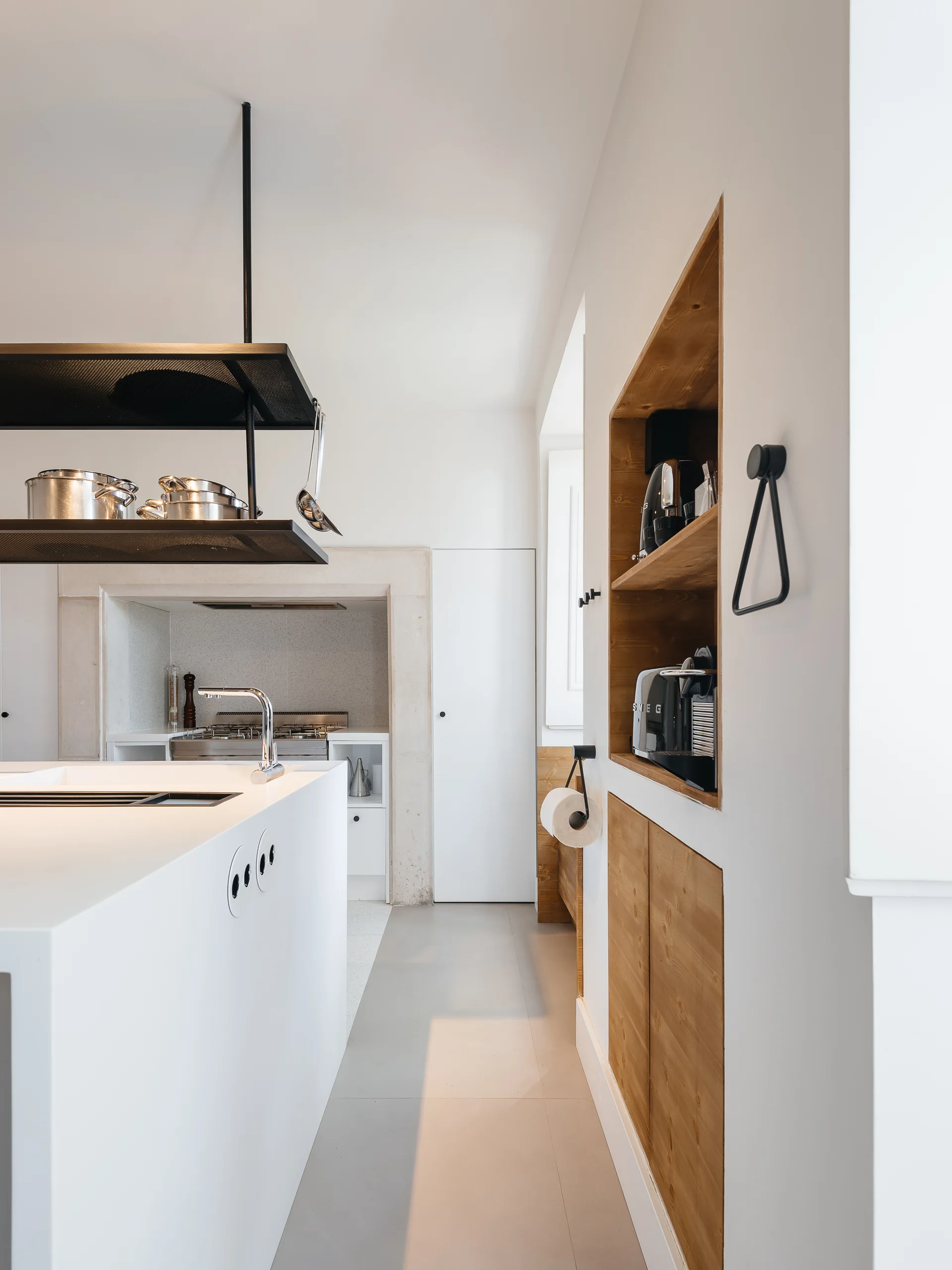 A minimalist white kitchen features a white island with a chrome faucet, an overhead black pot rack, and a recessed wood-toned wall niche.