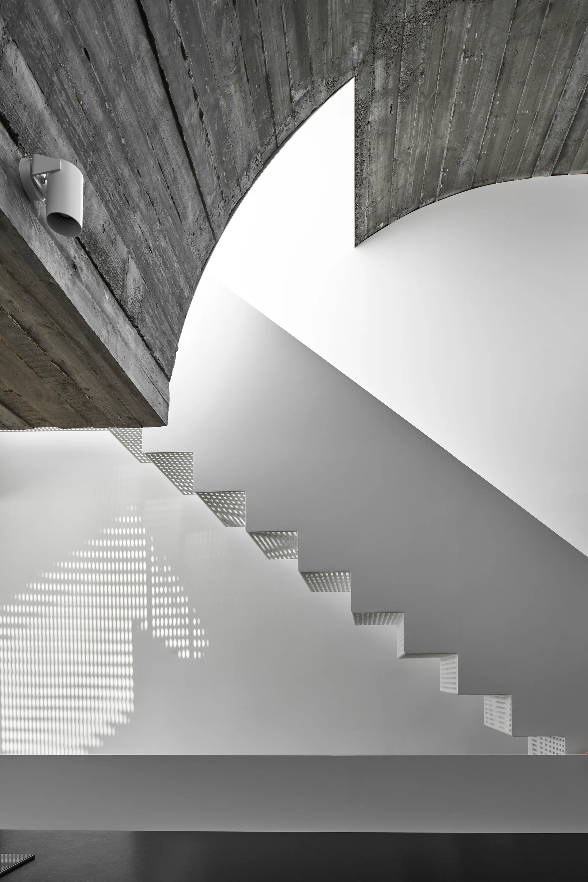 A minimalist white floating staircase with perforated metal treads casts shadows on a white wall beneath a curved textured concrete ceiling.